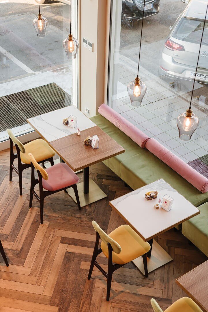 Ein modernes Café mit Holztischen, bunten Stühlen und Hängelampen. Der Raum hat große Fenster, die natürliches Licht hereinlassen, und eine gepolsterte Bank an der Wand. Draußen auf der Straße sind Autos zu sehen.
