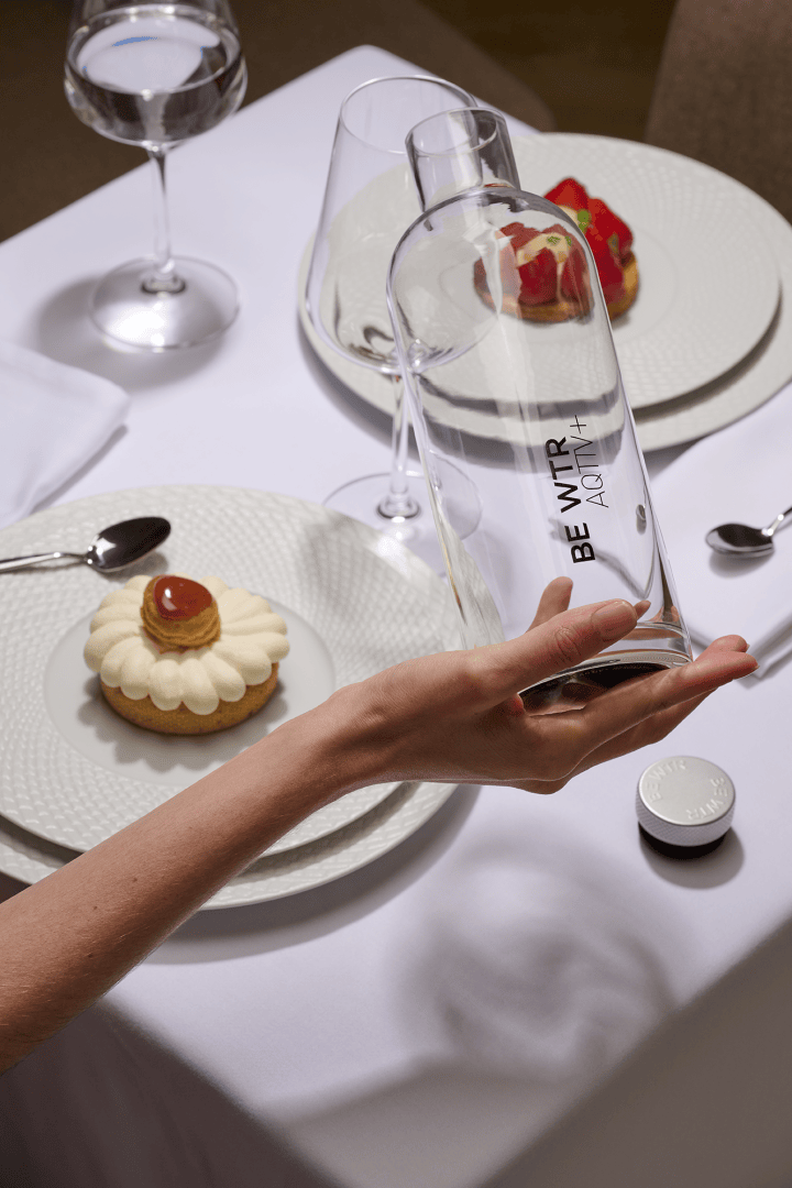 Une main tient une bouteille d'eau claire portant l'inscription "BEB WTR AQUA 14" au-dessus d'une table de salle à manger nappée de blanc, avec deux assiettes contenant des desserts élégants, des verres à vin, des couverts en argent et un verre d'eau.