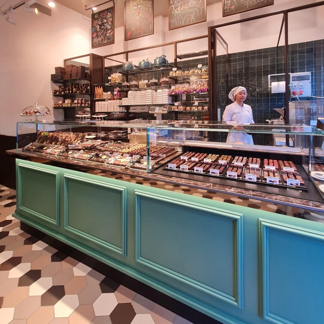 Eine Bäckerei oder ein Schokoladengeschäft mit einer Vitrine, die mit verschiedenen Backwaren und Pralinen gefüllt ist. Eine Mitarbeiterin in weißer Uniform und mit Hut steht hinter dem Tresen. Im Hintergrund sind Regale mit Gläsern, Schachteln und Dekorationen zu sehen.