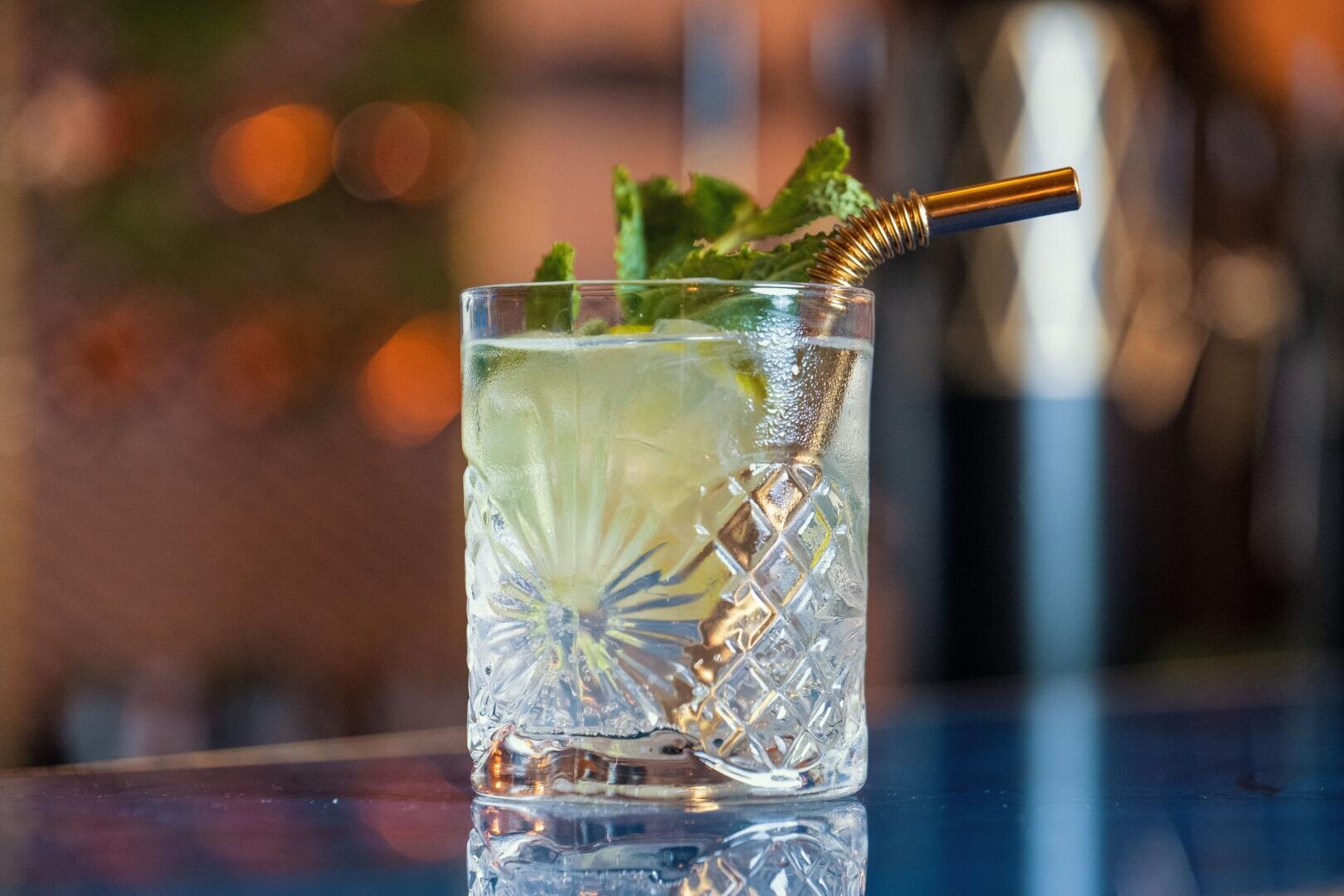 Ein klares Glas mit verschnörkeltem Muster enthält einen hellgrünen Cocktail, der mit frischen Minzblättern garniert und mit einem Metallstrohhalm serviert wird, auf einer spiegelnden blauen Oberfläche mit verschwommenem Hintergrund.