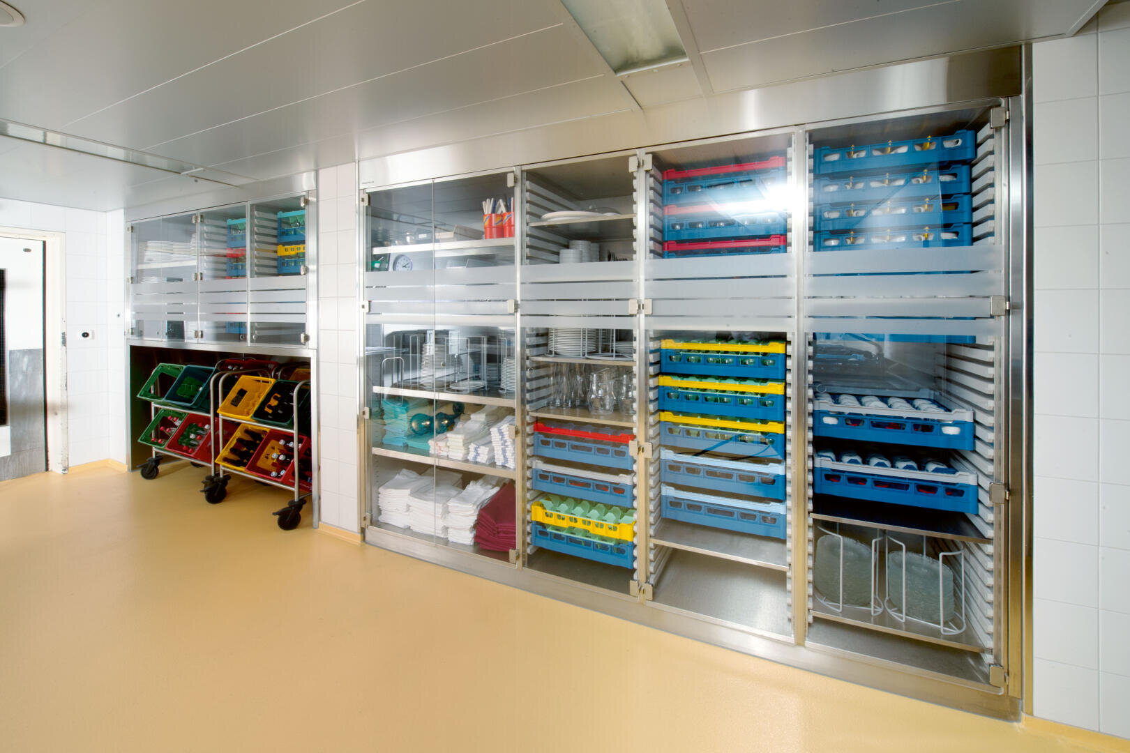 Une salle de stockage propre et organisée avec des étagères métalliques contenant des plateaux, des bacs et des fournitures de cuisine derrière des portes vitrées ; des chariots à roulettes avec des bacs colorés se trouvent sur la gauche. La pièce a des murs en carrelage blanc et un sol jaune.