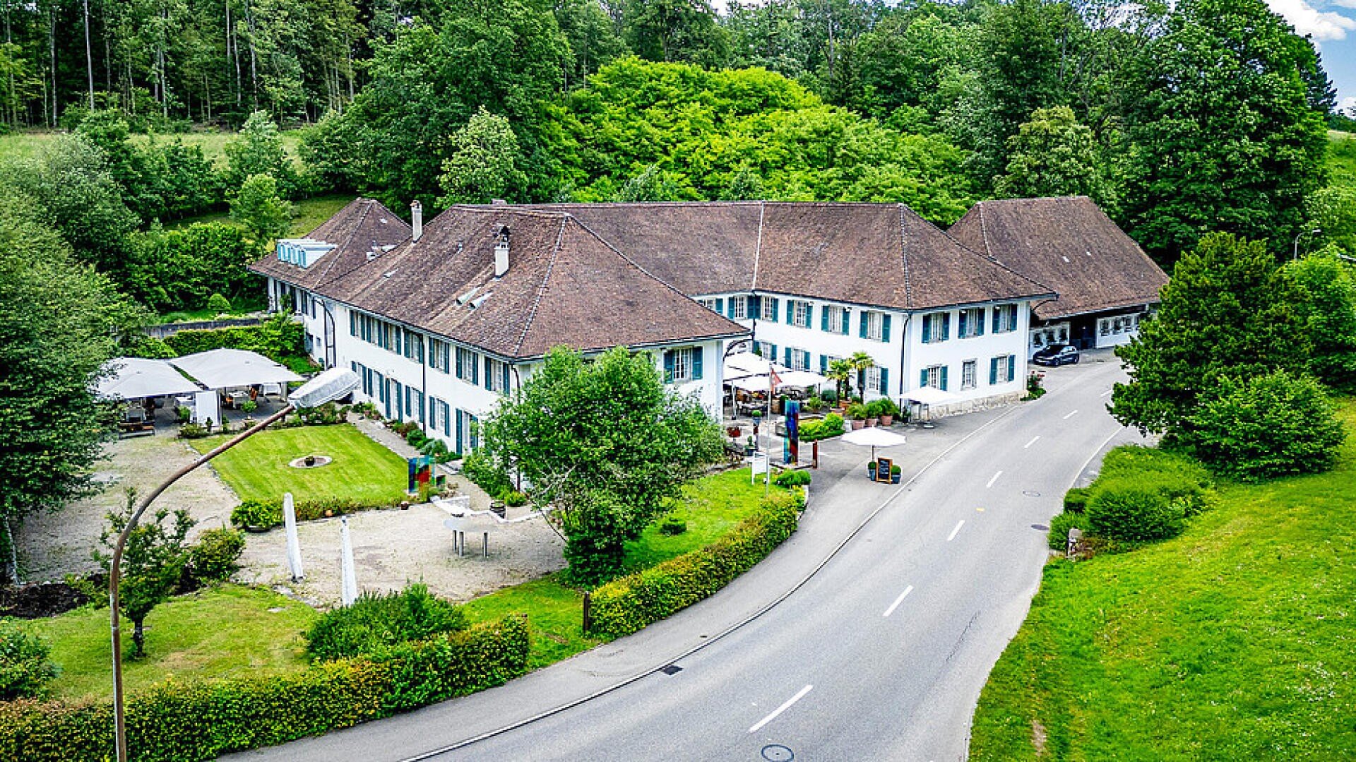 Luftaufnahme eines großen weißen Gebäudes mit braunem Ziegeldach, das von grünen Bäumen und Rasen umgeben ist und an einer kurvigen Straße liegt. Sitzgelegenheiten im Freien und Sonnenschirme sind in der Nähe des Eingangs zu sehen.