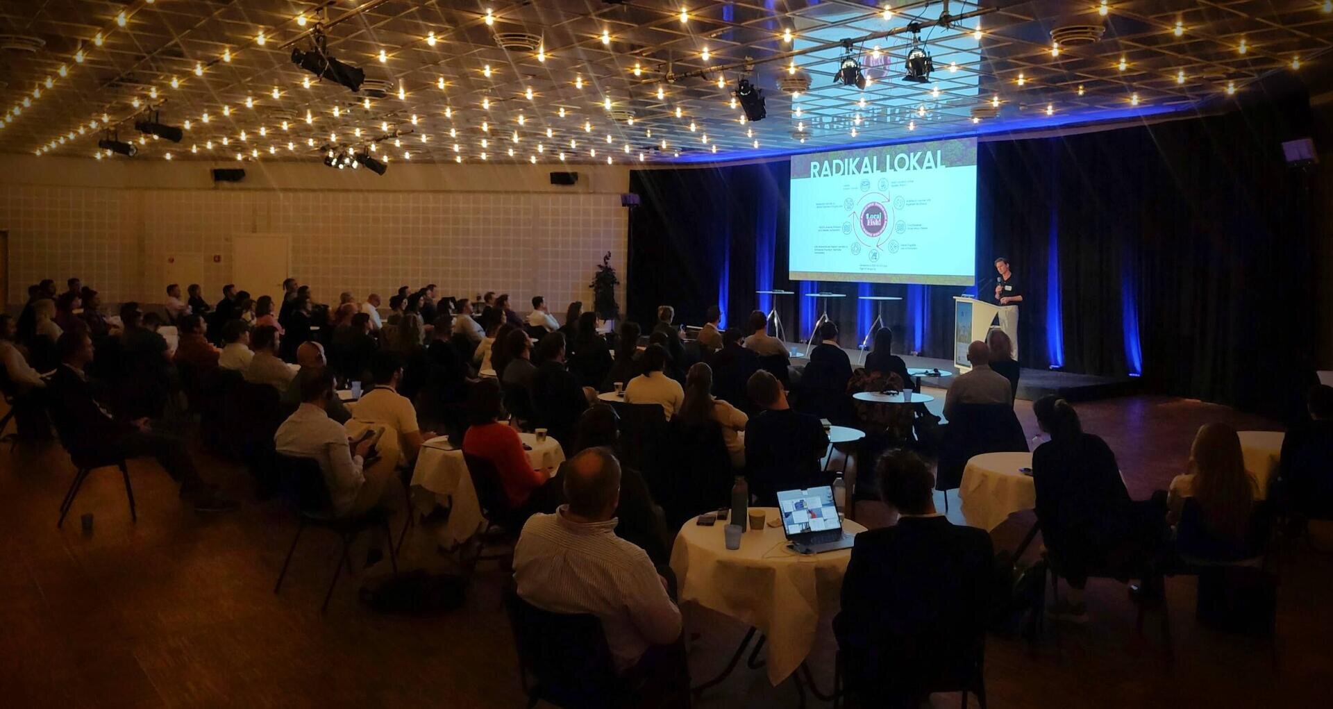 Ein Redner spricht in einem schwach beleuchteten Konferenzraum zu einem großen Publikum, wobei eine Folie mit dem Titel RADIKALLOKAL auf eine Leinwand projiziert wird. Die Teilnehmer sitzen an runden Tischen und hören aufmerksam zu.