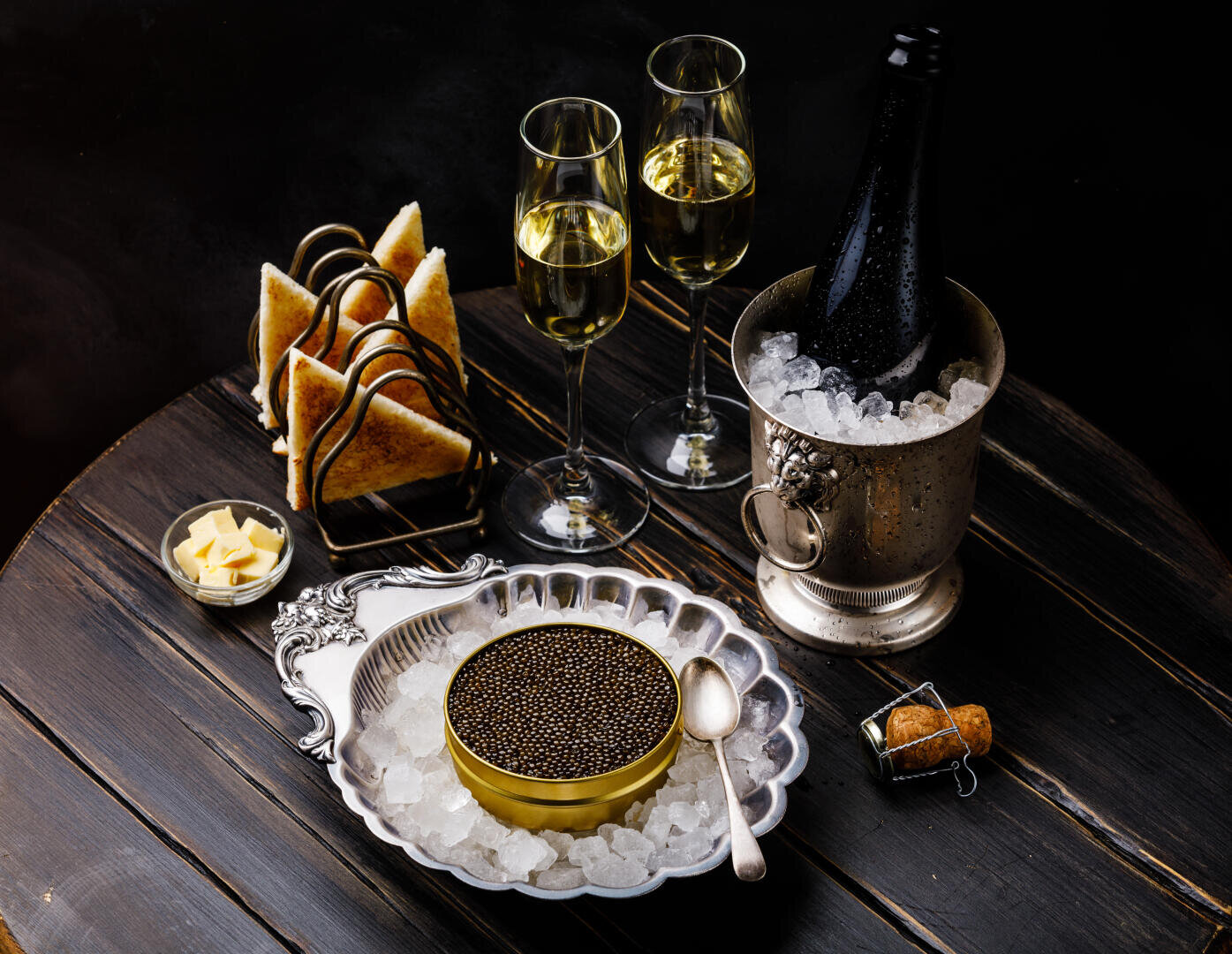 Une boîte de caviar noir sur glace avec une cuillère, deux coupes de champagne, une bouteille dans un seau à glace, des tranches de pain grillé et des cubes de beurre sur une table ronde en bois foncé.