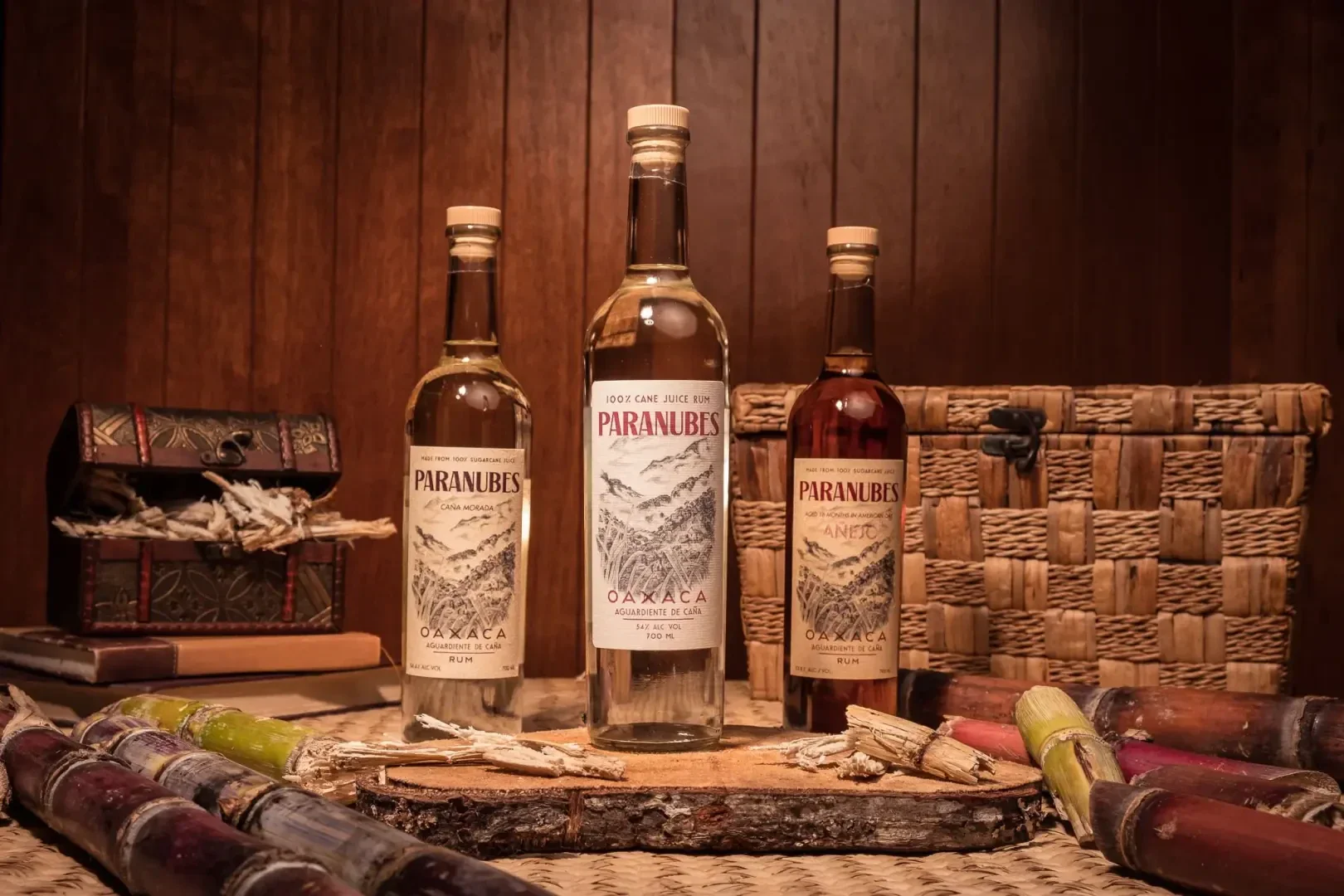 Trois bouteilles de rhum Paranubes sont posées sur une surface en bois avec des tiges de canne à sucre, des livres, un coffre et un panier tressé en arrière-plan, le tout devant un mur en boiseries.
