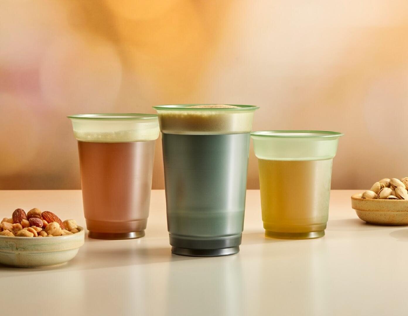 Drei Getränke in Milchglasbechern, jeder mit einer Schaumschicht, stehen auf einer hellen Fläche. Zwei kleine Schalen mit verschiedenen Nüssen und Pistazien stehen neben den Bechern. Der Hintergrund ist sanft verschwommen.