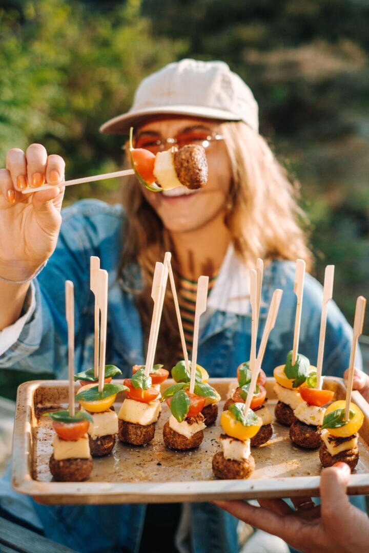 Eine lächelnde Frau mit Mütze und Sonnenbrille hält ein Tablett mit bunten Spießen mit Bits for Kids, Käse, Tomaten und Basilikum in der Hand und nimmt einen davon im Freien in der Sonne auf.
