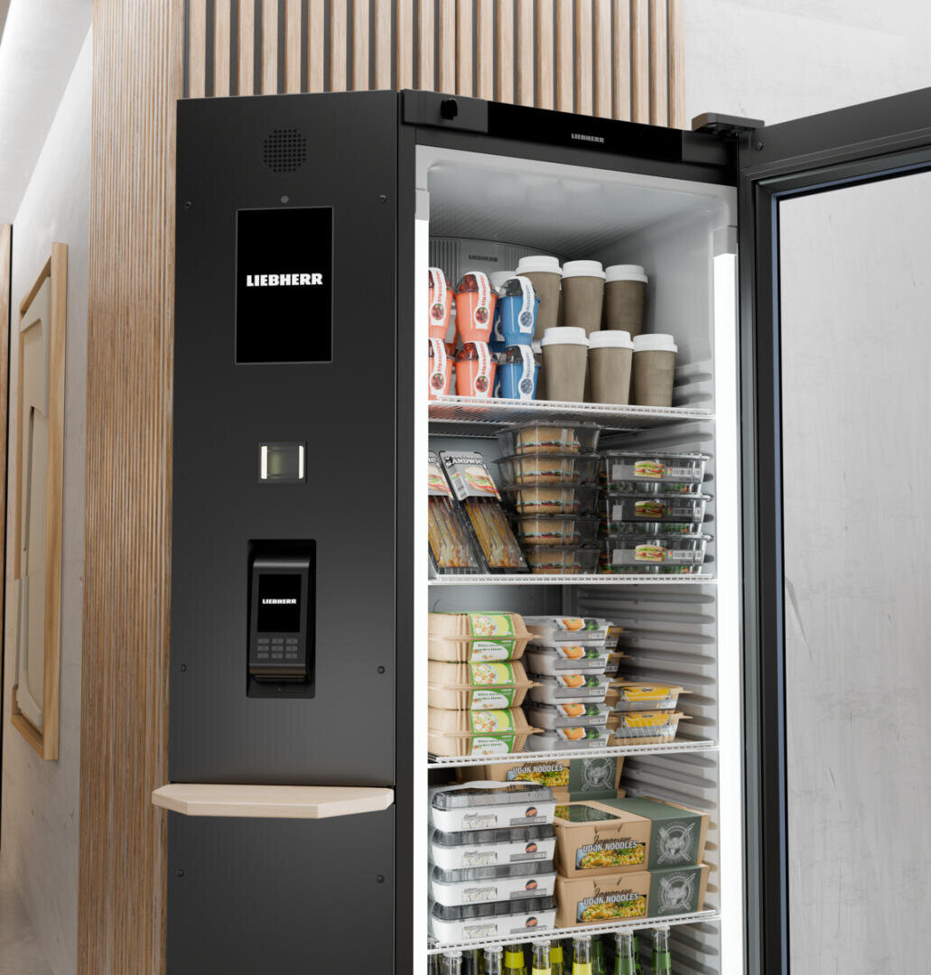 Un réfrigérateur automatique avec une porte vitrée propose différents repas emballés, des boissons, des gobelets à café empilables et des œufs. L'appareil est de la marque Liebherr et dispose d'un système de paiement par écran tactile.
