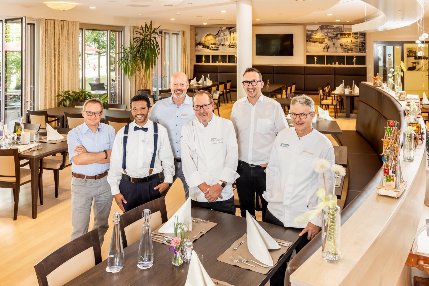 Sechs Männer, davon drei in Kochmänteln und drei in eleganter Freizeitkleidung, stehen lächelnd in einem hellen, modernen Restaurant mit ordentlich gedeckten Tischen und zeitgenössischem Dekor.