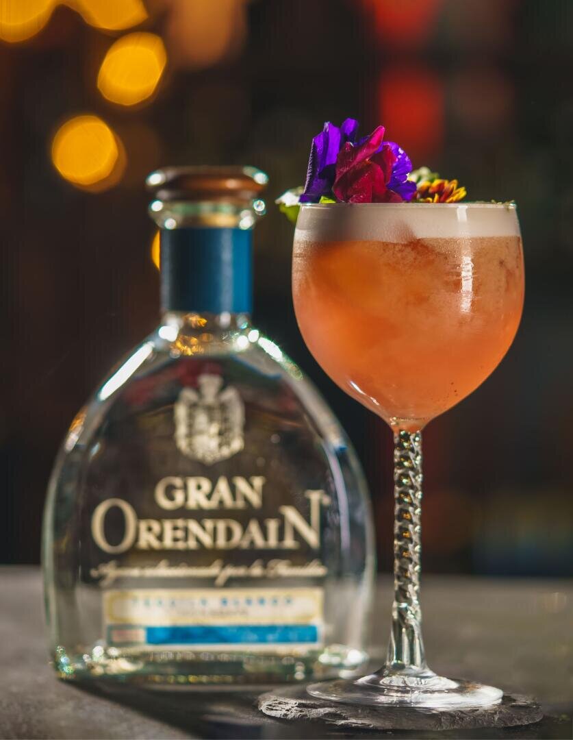 Eine Flasche Gran Orendain Tequila steht neben einem Cocktail in einem dekorativen Glas, garniert mit lebhaften essbaren Blumen, vor einem sanft verschwommenen Hintergrund mit warmen Lichtern.