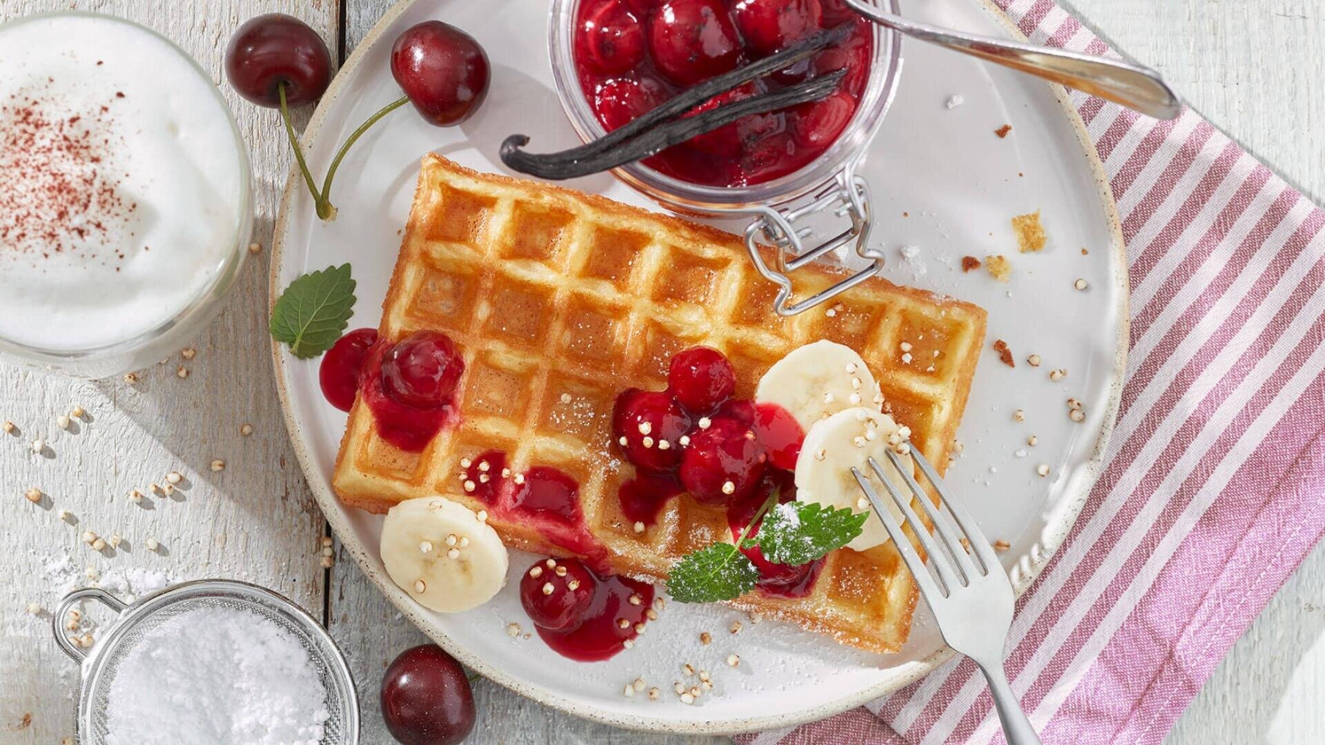 Une assiette avec une gaufre dorée garnie de sauce aux cerises, de rondelles de banane et de menthe, à côté d'un pot de compote de cerises, de cerises fraîches, d'une tasse de lait mousseux et d'un bol de sucre en poudre sur une serviette rayée.