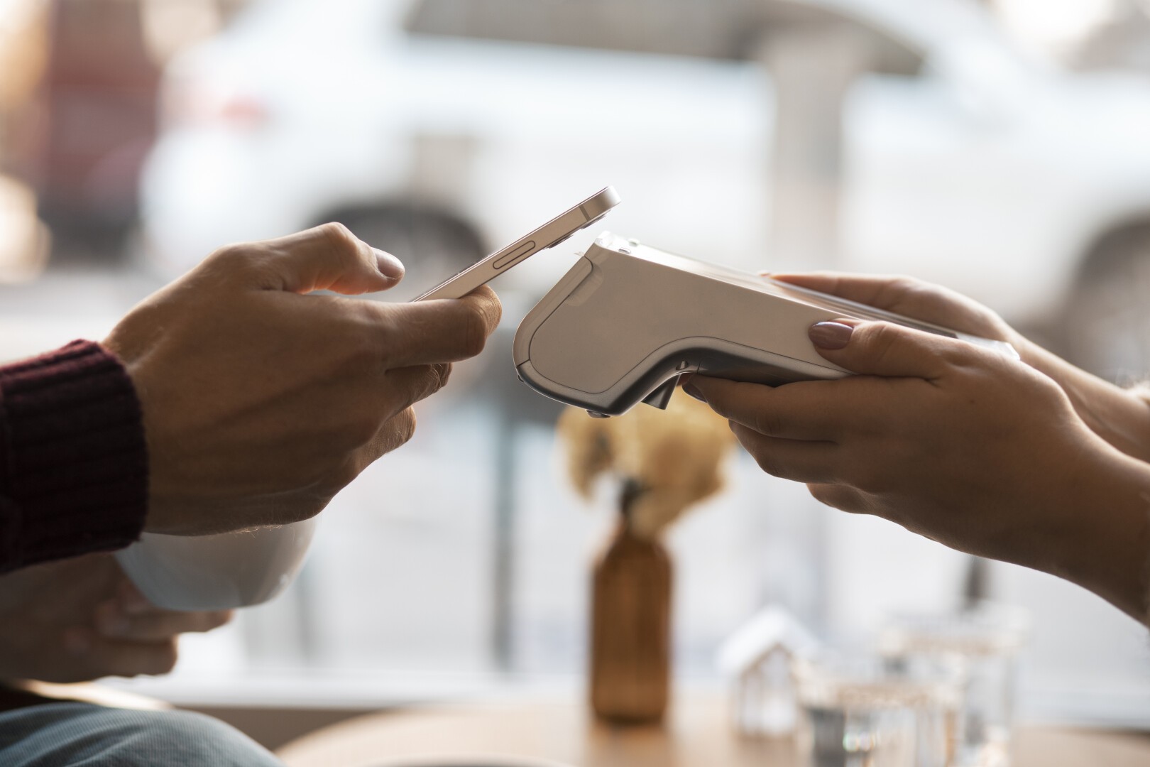 Zwei Personen bezahlen kontaktlos mit einem Smartphone und einem Kartenlesegerät. Eine Person hält das Smartphone, die andere das Terminal. Der Hintergrund ist unscharf.