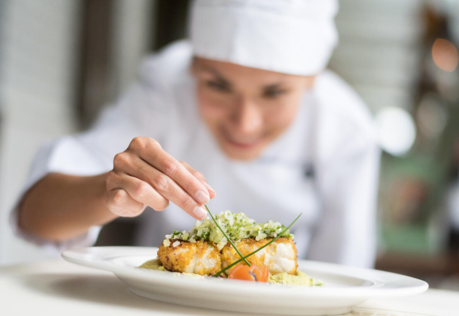 Ein Koch in weißer Uniform und Mütze garniert sorgfältig ein Gericht mit frischen Kräutern und achtet dabei auf die Details. Das Gericht besteht aus paniertem Fisch und Gemüse und wird auf einem weißen Teller präsentiert.