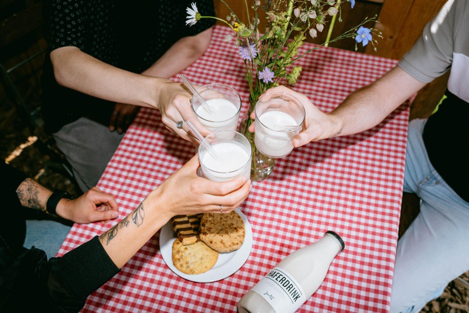 Vier Personen stoßen mit Milchgläsern an einem rot karierten Tischtuch an. Auf dem Tisch stehen ein Teller mit Keksen, eine Flasche mit der Aufschrift MYDRINK und eine Vase mit Wildblumen.