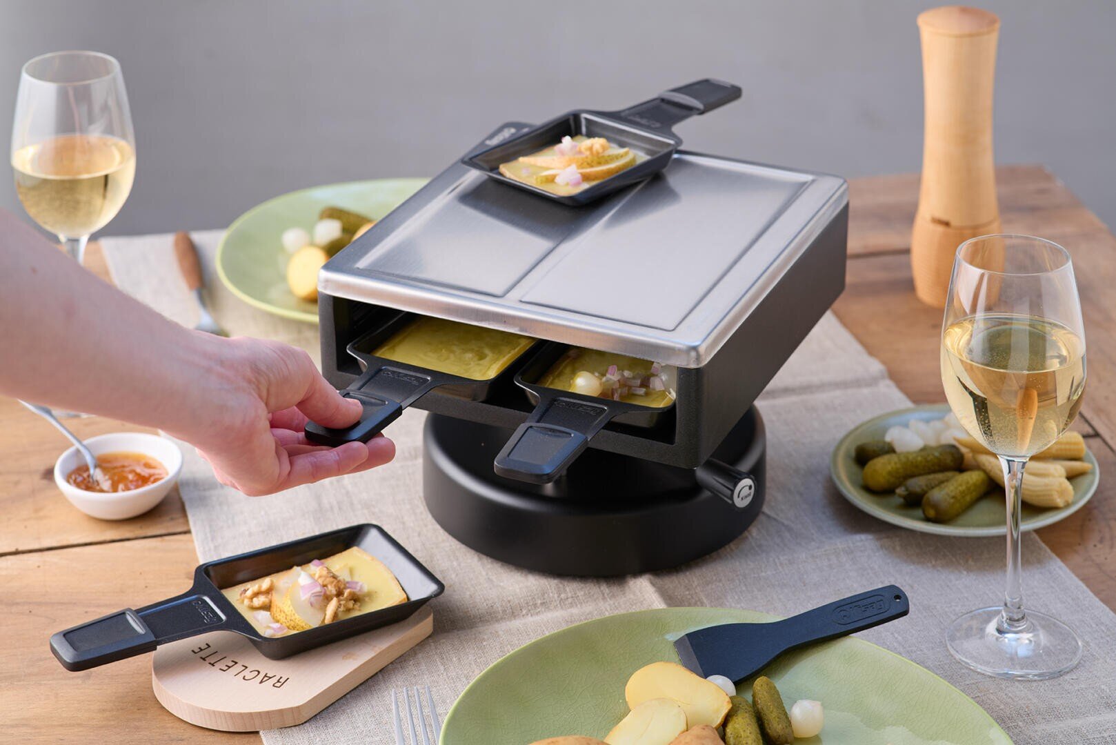 Eine Hand stellt kleine Pfännchen mit geschmolzenem Käse und Toppings in einen Tisch-Raclette-Grill. Auf einem Holztisch sind Teller mit Kartoffeln, Essiggurken, Zwiebeln, Weingläsern und Gewürzen angeordnet.