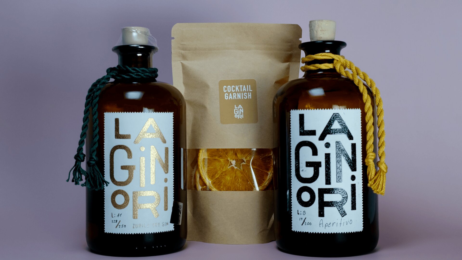 Zwei braune Glasflaschen mit der Aufschrift LA GINORI und dekorativen Kordeln sowie ein brauner Papierbeutel mit der Aufschrift Cocktail Garnish, der getrocknete Orangenscheiben enthält, sind vor einem blassvioletten Hintergrund ausgestellt.