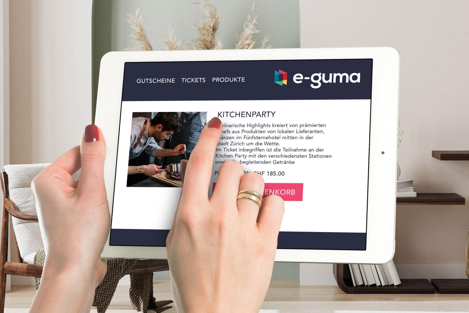 Eine Person hält und benutzt ein weißes Tablet und betrachtet eine Webseite über eine Küchenparty auf der e-guma-Website in deutscher Sprache mit einem Produktbild und Veranstaltungsdetails auf dem Bildschirm.