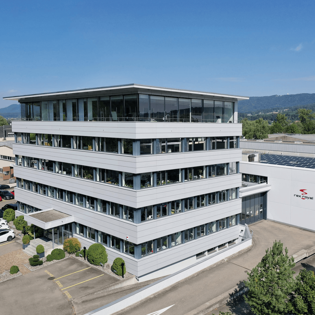 Un immeuble de bureaux moderne de plusieurs étages, avec de grandes baies vitrées et un toit plat, entouré d'espaces verts et d'un parking, sous un ciel bleu limpide. Un logo portant l'inscription Rex Royal est visible sur un bâtiment adjacent.