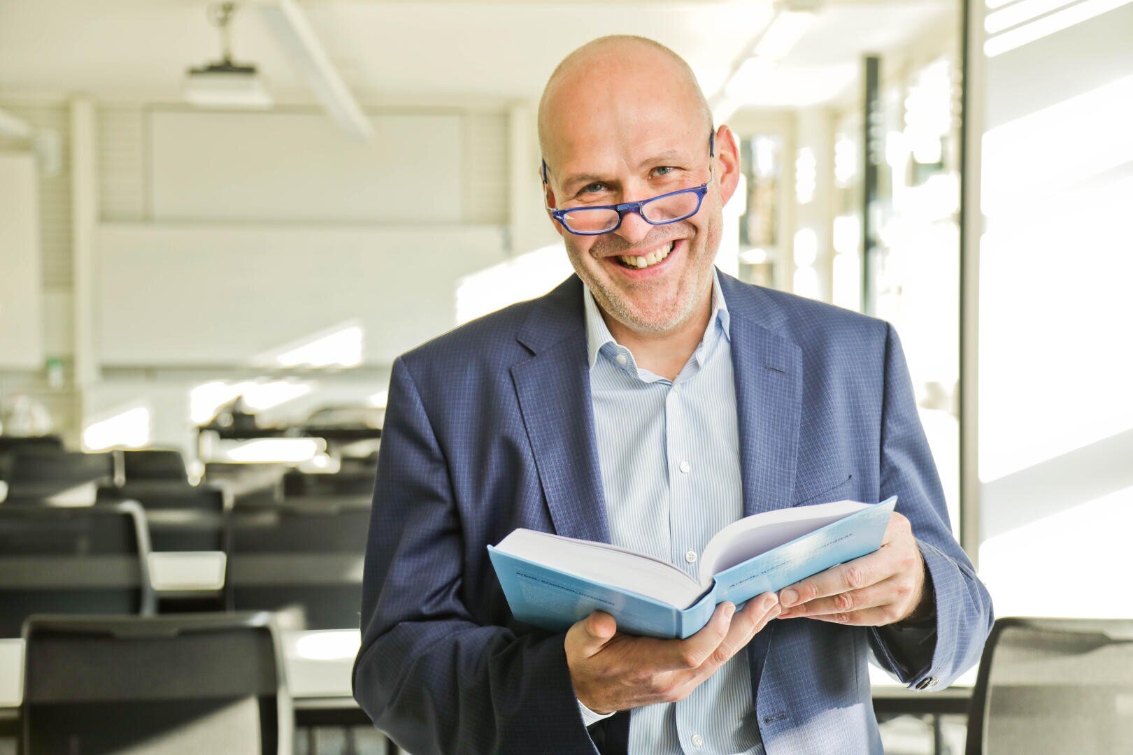 Ein lächelnder Mann mit Glatze, blauem Anzug und Brille hält ein aufgeschlagenes Buch in einem hellen, leeren Klassenzimmer mit Reihen von Stühlen und Tischen im Hintergrund.