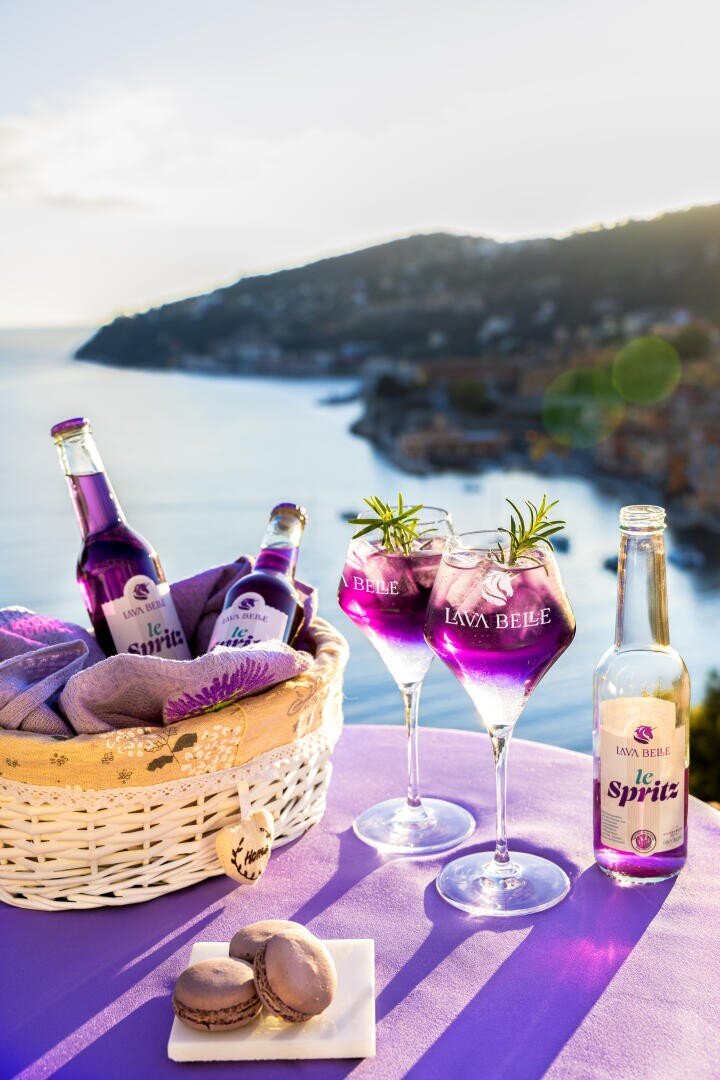 Un panier de boissons violettes, deux verres garnis de romarin et une bouteille sont posés sur une nappe de lavande avec vue sur un paysage côtier au coucher du soleil, précédés de macarons sur une assiette blanche.