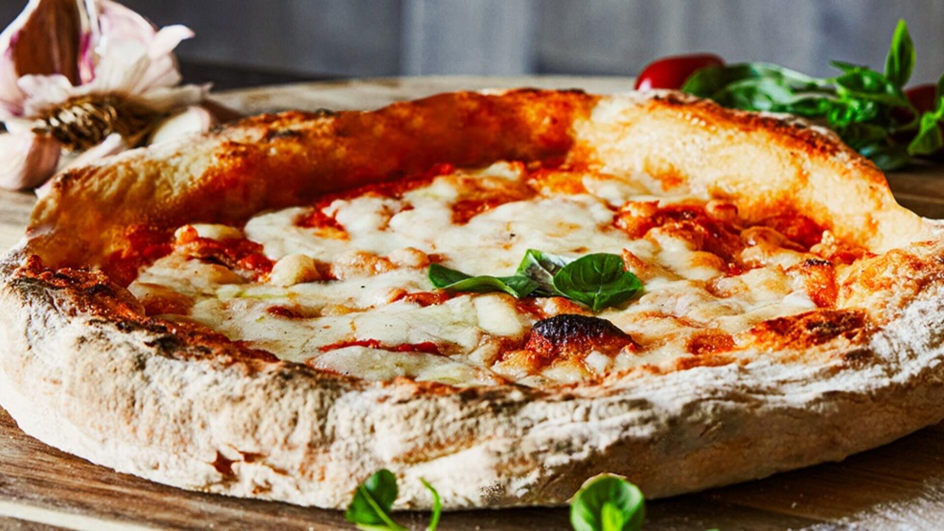 Nahaufnahme einer frisch gebackenen Pizza mit einer dicken, goldbraunen Kruste, geschmolzenem Käse, Tomatensoße und frischen Basilikumblättern auf einer Holzunterlage mit Kräutern und Knoblauch im Hintergrund.