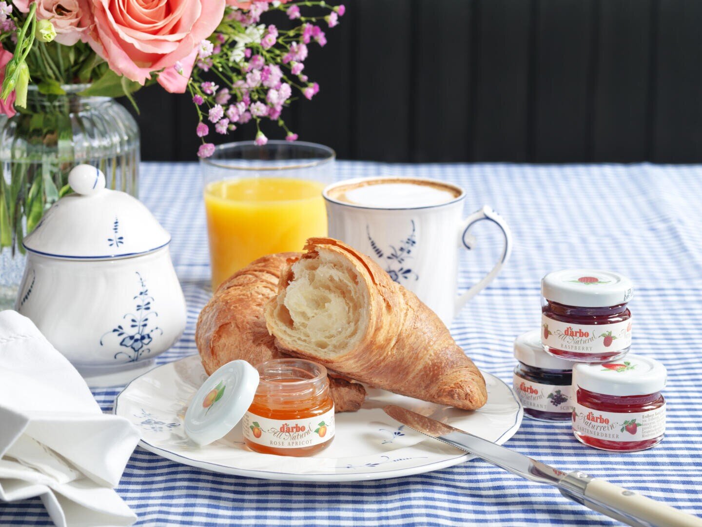 Ein Croissant auf einem Teller mit einem offenen Glas Aprikosenmarmelade, Minigläsern mit verschiedenen Fruchtmarmeladen, einem Messer, einer Tasse Kaffee, einem Glas Orangensaft und einer Vase mit Blumen auf einer blau karierten Tischdecke.