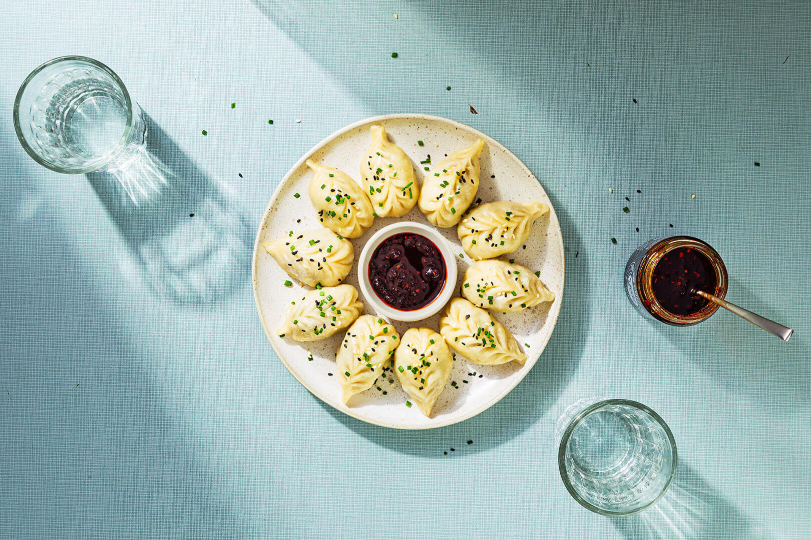 Une assiette de boulettes bien disposées avec une garniture verte entoure un petit bol de sauce dip foncée ; trois verres d'eau et un verre d'huile pimentée sont posés à côté sur un plateau de table bleu clair.