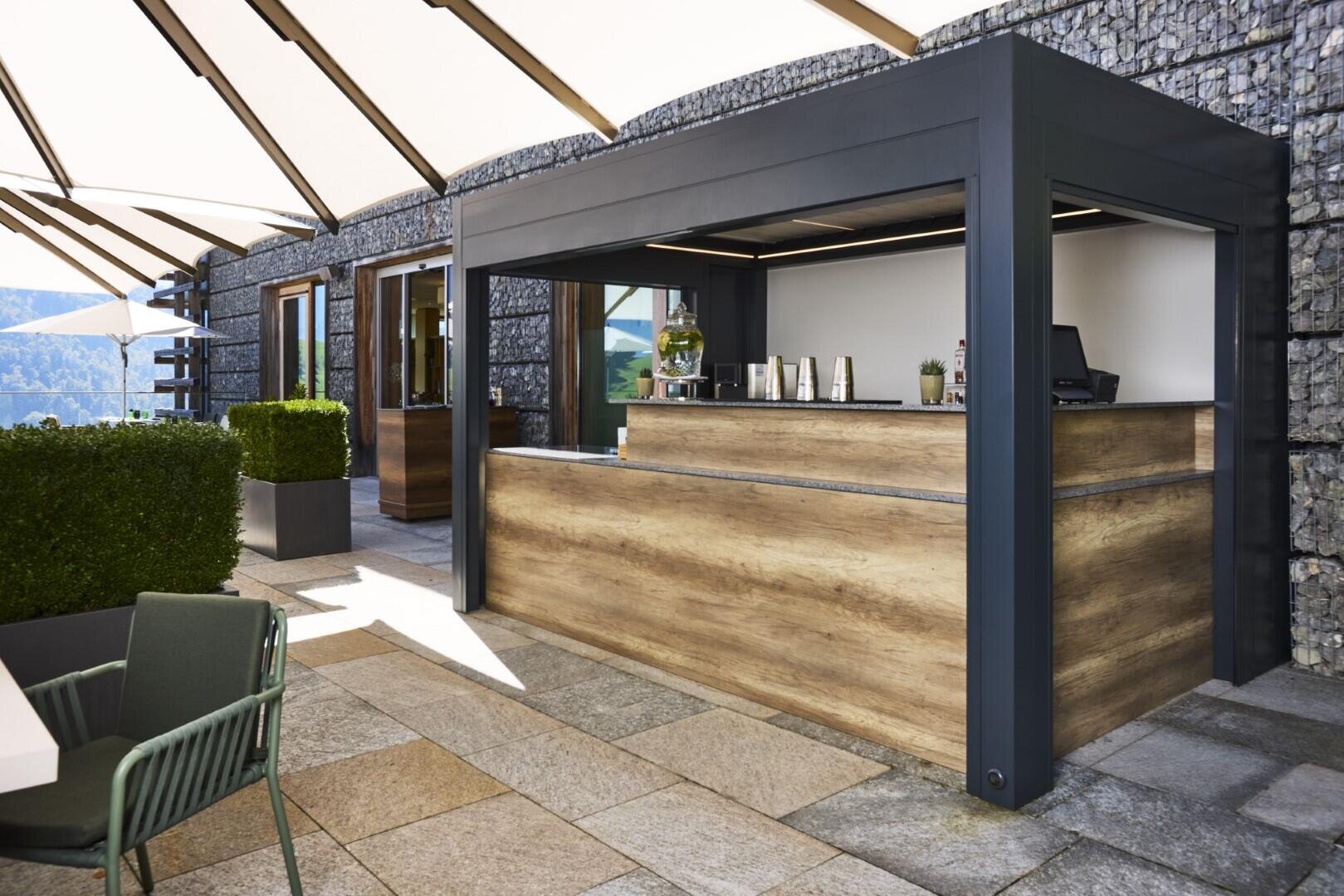 Ein modernes Café mit holzgetäfeltem Tresen unter einem Vordach auf einer Steinterrasse mit grünen Sträuchern und Stühlen in der Nähe. Steinmauern und Sonnenschirme spenden Schatten und sorgen für eine malerische, entspannte Atmosphäre.