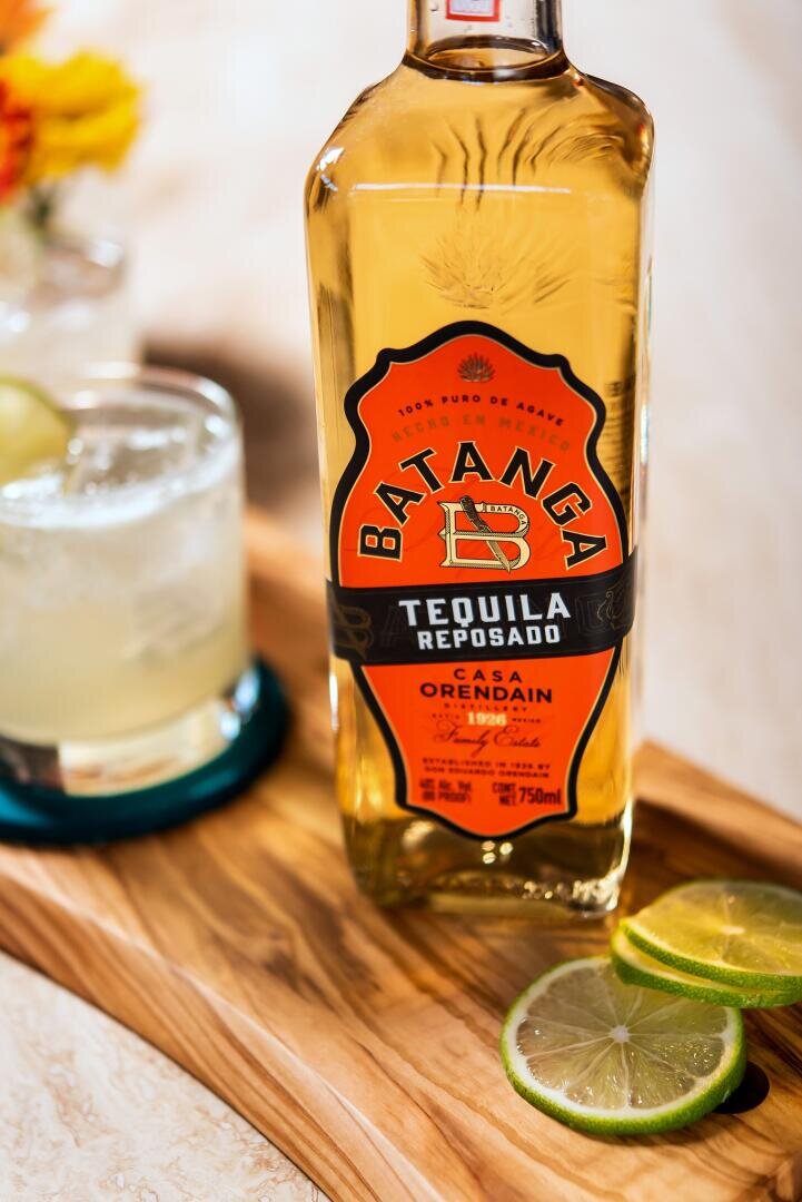 Eine Flasche Batanga Reposado Tequila steht auf einem Holztablett neben einer aufgeschnittenen Limette und einem Cocktailglas mit Limettengarnitur, auf einer hellen Fläche mit verschwommenen Blumen im Hintergrund.