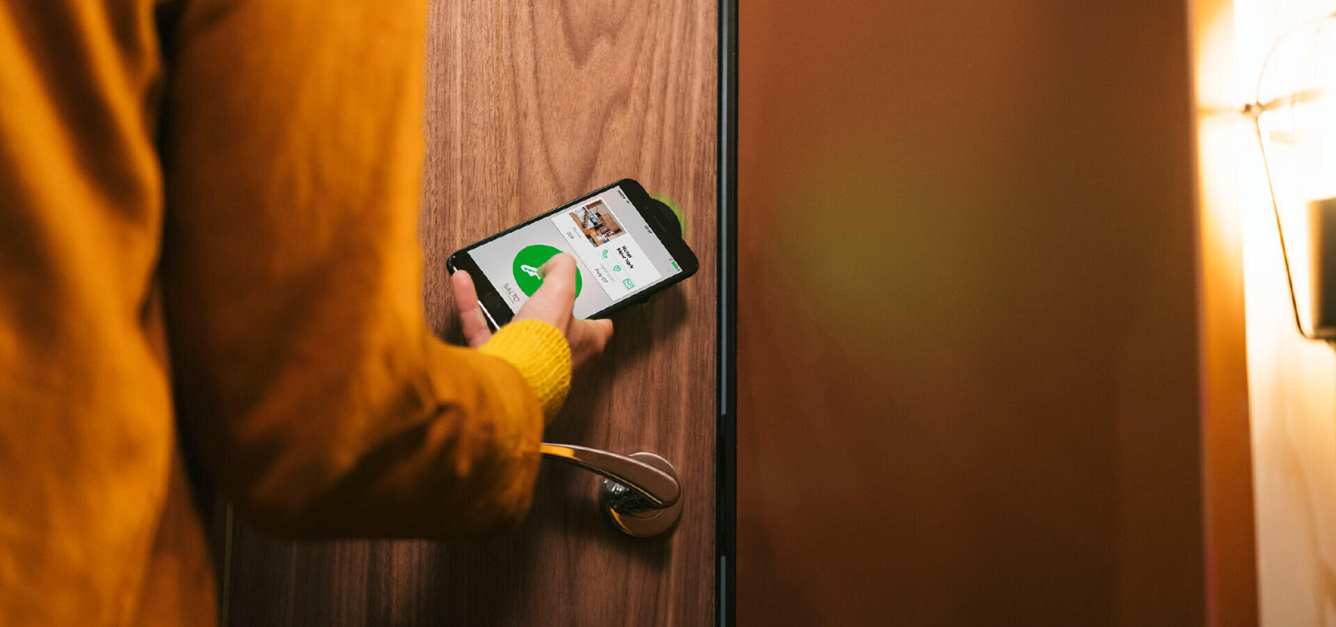 Une personne vêtue d'un pull jaune utilise un smartphone avec une application pour déverrouiller une porte en bois, en tenant le téléphone près de la poignée.
