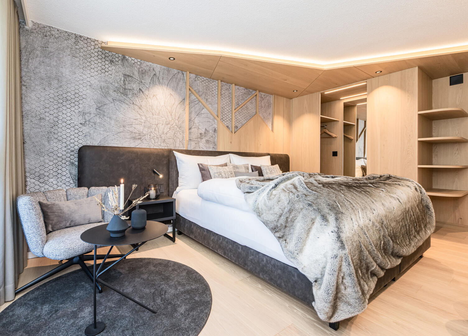 Modernes Schlafzimmer mit großem Bett, grauem Polsterkopfteil, Felldecke, Sessel, rundem Couchtisch und eingebauten Holzregalen. Sanfte Beleuchtung und helle Holzakzente schaffen eine gemütliche, stilvolle Atmosphäre.
