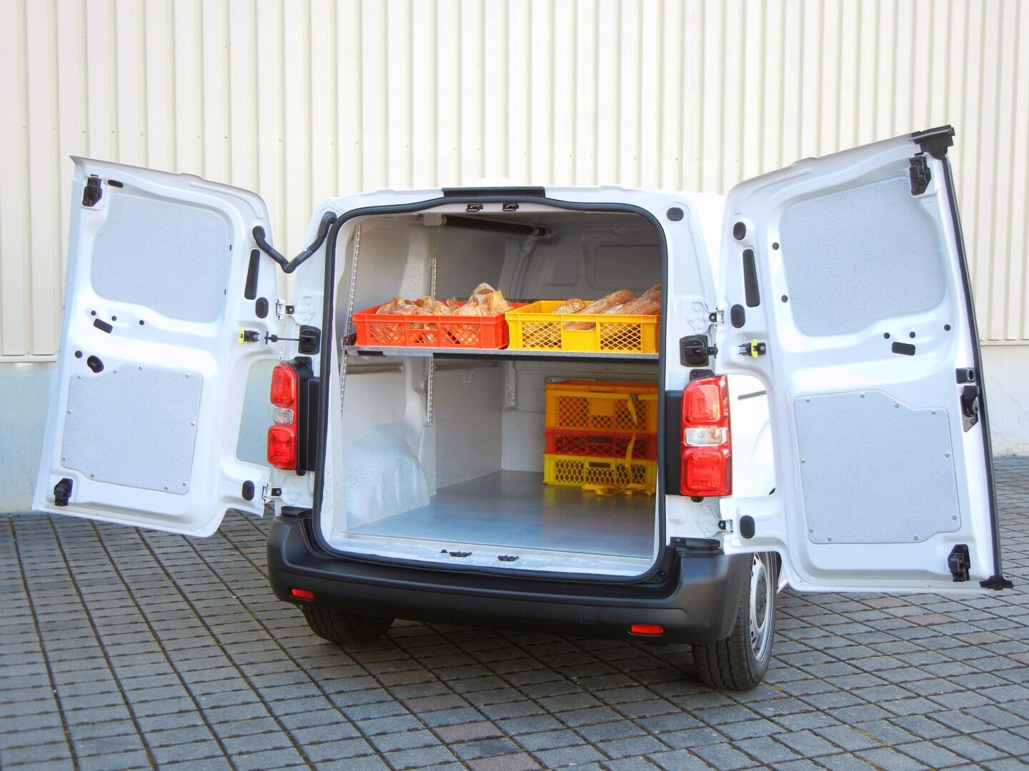 Une camionnette blanche dont les portes arrière sont ouvertes et qui contient des caisses jaunes et orange de pain et de petits pains. La camionnette est garée sur une surface pavée à côté d'un bâtiment beige.