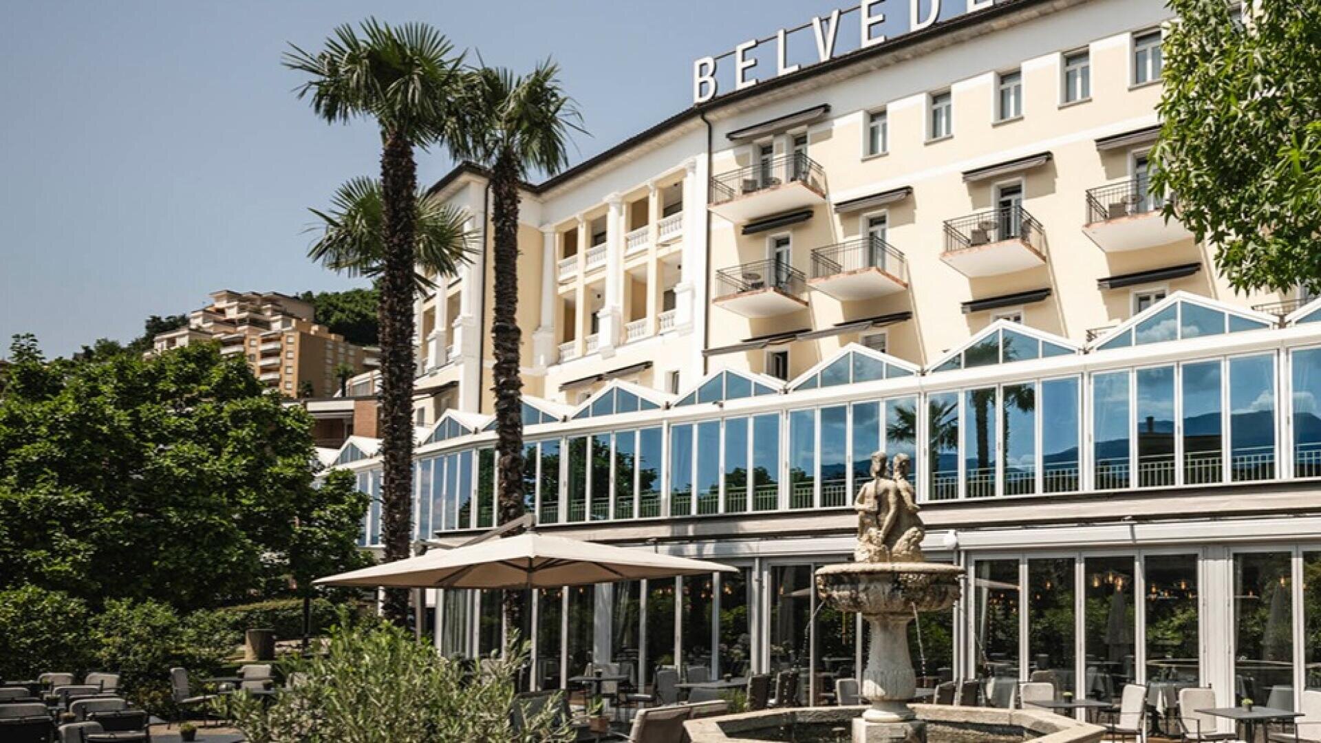Ein prächtiges Hotelgebäude mit Balkonen und großen Glasfenstern, umgeben von Palmen und Grünanlagen. Davor befindet sich ein Innenhof mit Sitzgelegenheiten im Freien und einem Steinbrunnen mit einer Statue.