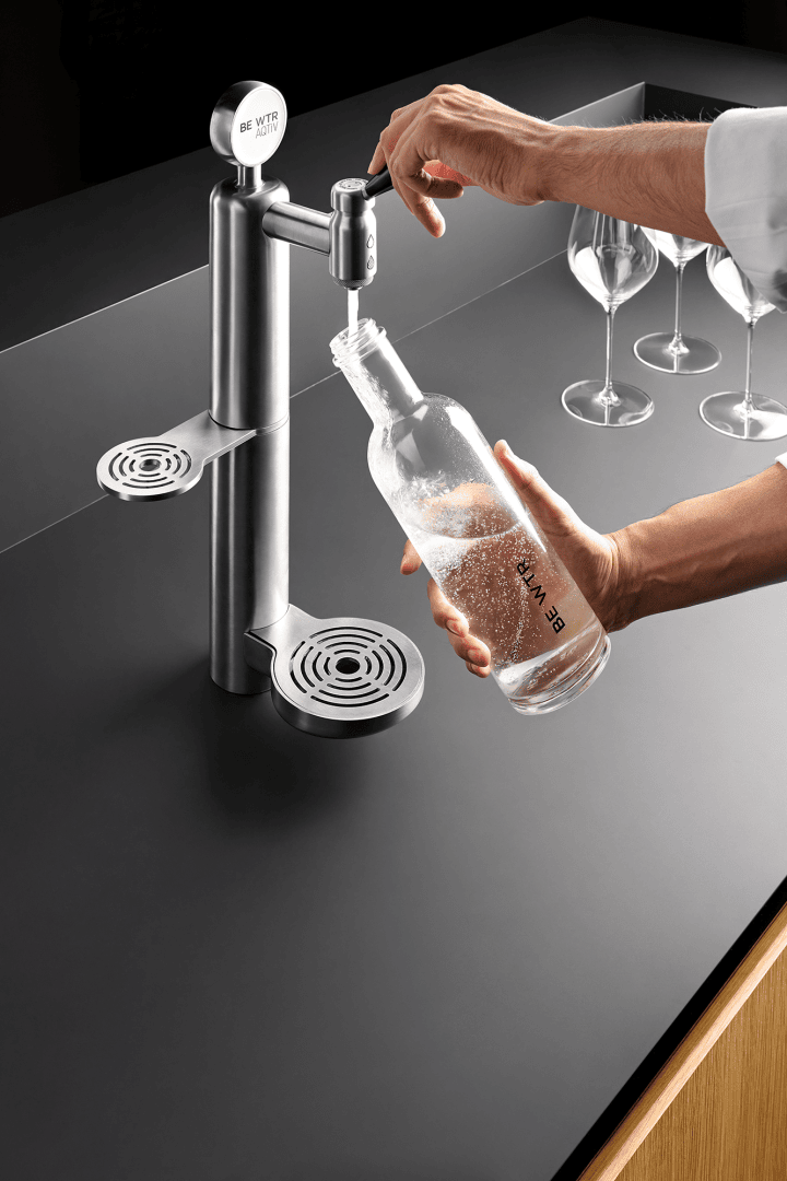 Une personne remplit une bouteille en verre d'eau gazeuse à partir d'un robinet moderne en acier inoxydable posé sur un plan de travail noir. En arrière-plan, on aperçoit trois verres à vin vides.