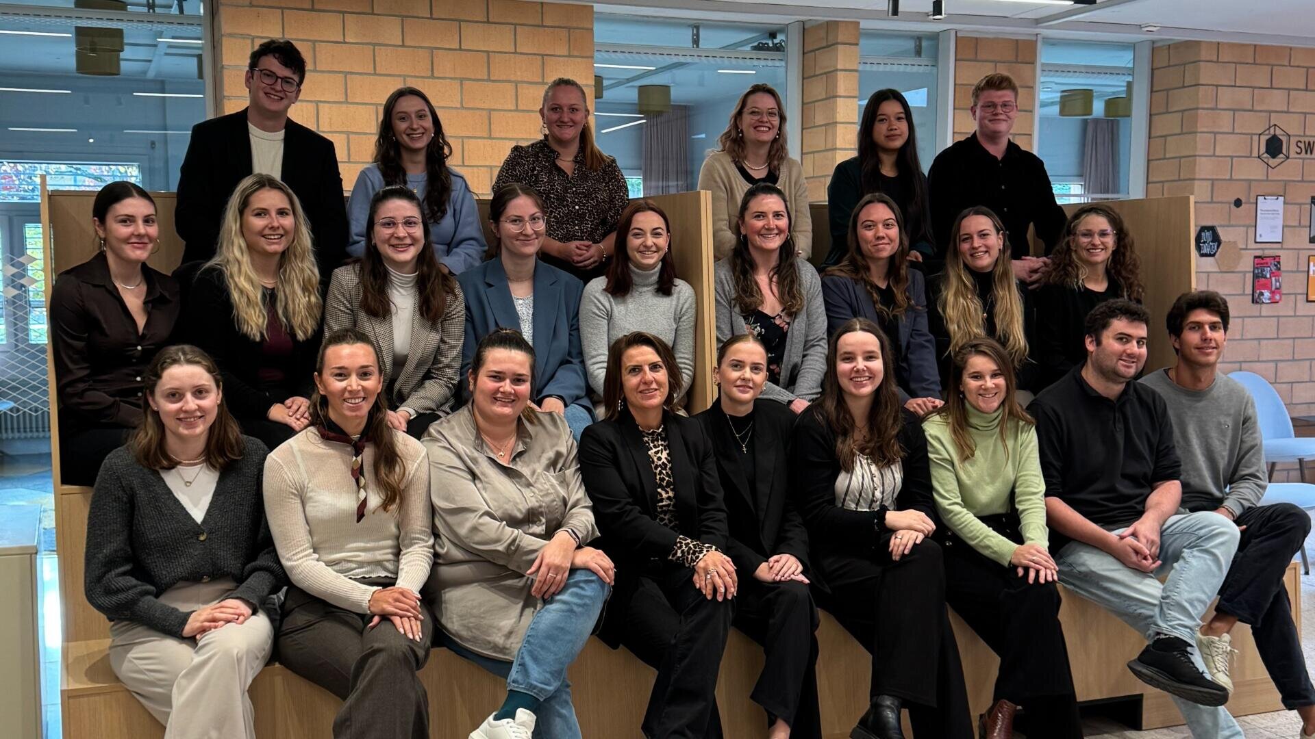 Eine Gruppe von 24 Personen, meist Frauen, sitzt und steht auf Holzbänken in einer modernen Inneneinrichtung und lächelt in die Kamera. Im Hintergrund sind Backsteinwände und große Fenster zu sehen.