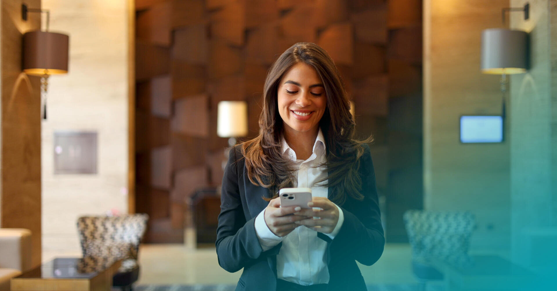 Une femme en tenue d'affaires sourit en regardant son smartphone dans un lobby moderne et élégant au décor contemporain et à l'éclairage doux.