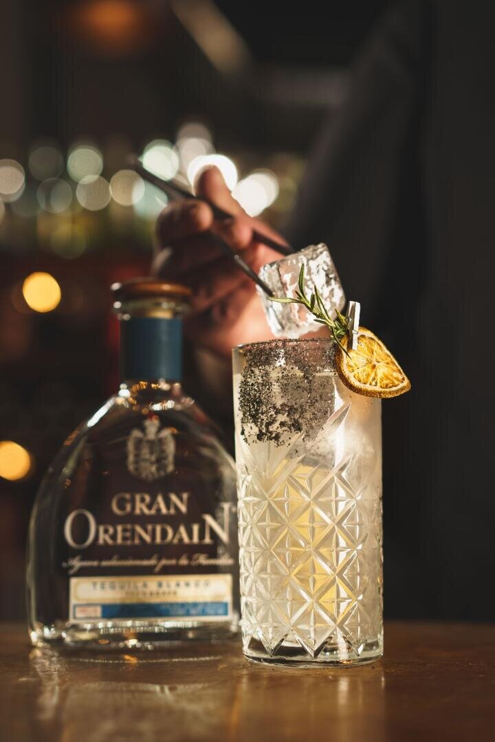 Eine Hand rührt einen Cocktail mit Eis in einem Kristallglas, garniert mit einer getrockneten Zitrusscheibe und Rosmarin, neben einer Flasche Gran Orendain Tequila auf einem Bartresen.