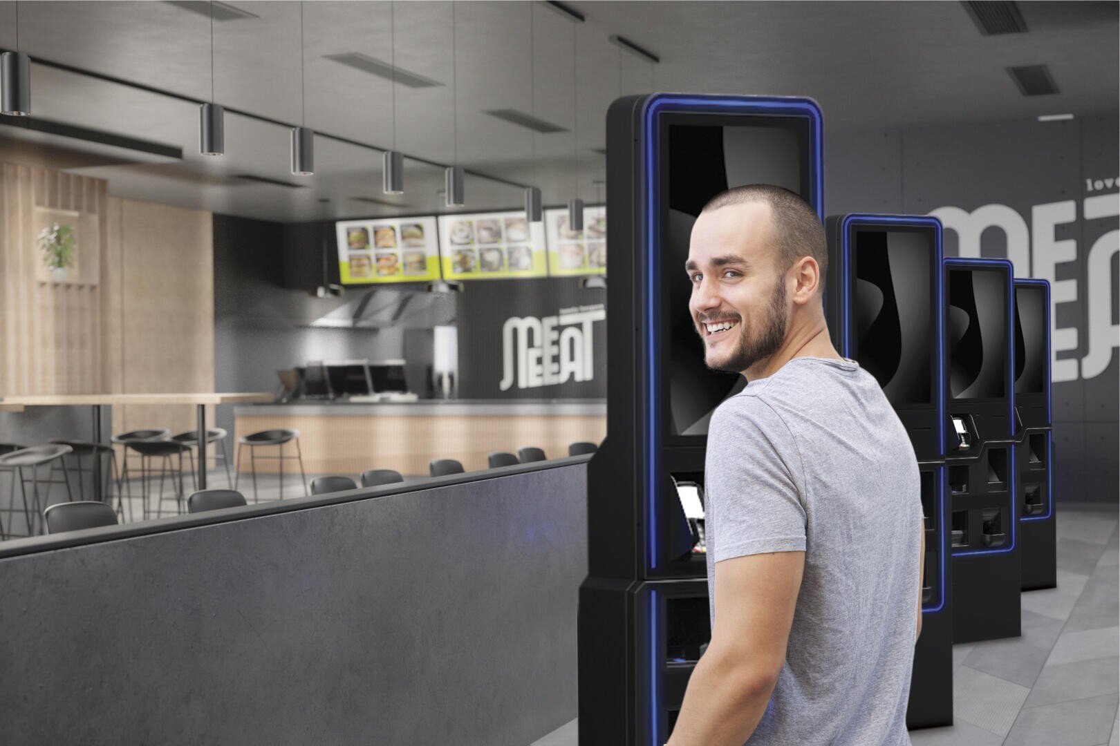 Un homme souriant vêtu d'un T-shirt gris se tient à un kiosque libre-service dans un fast-food moderne et vide, avec un décor minimaliste et des tableaux de menus en arrière-plan.