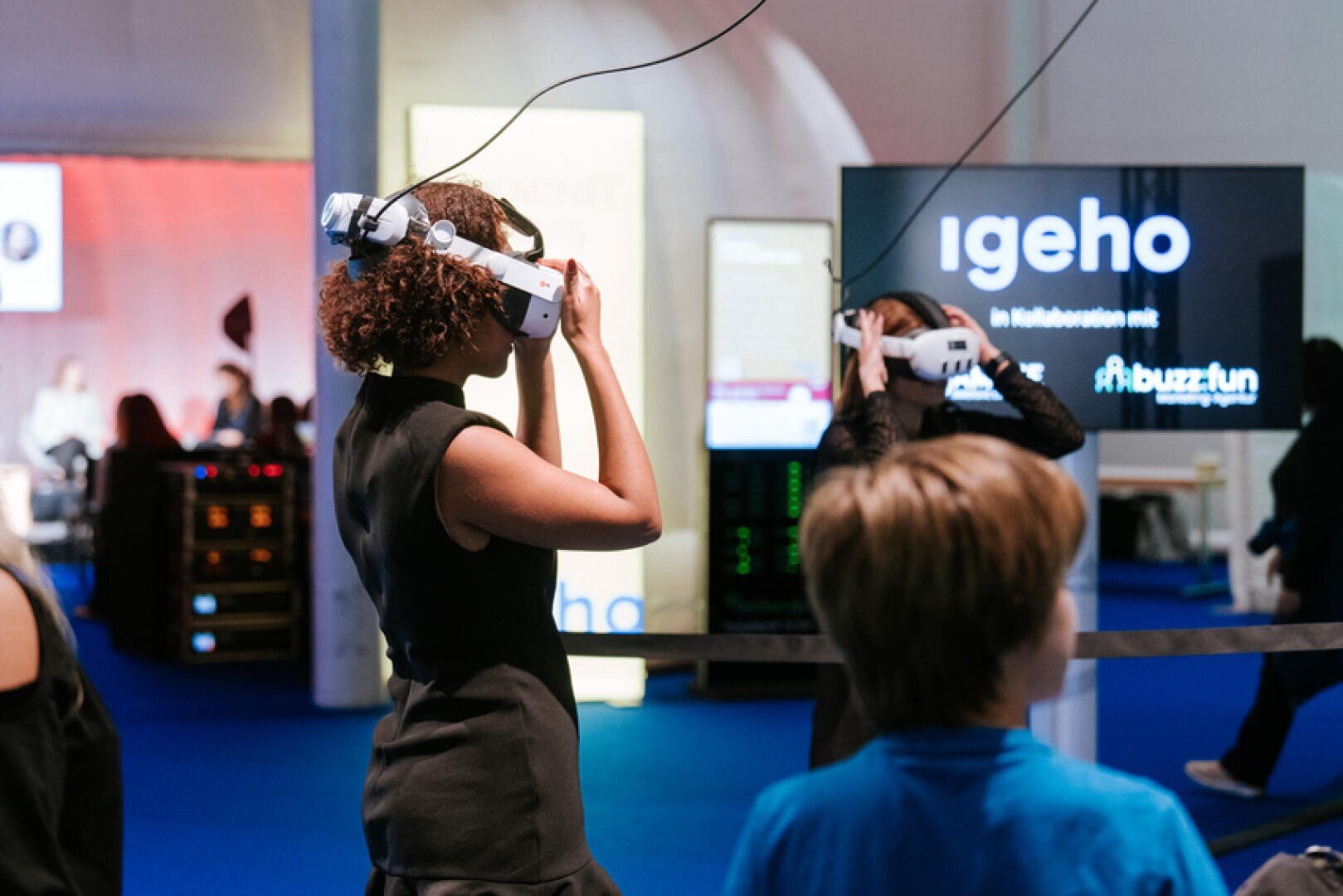 Zwei Personen tragen bei einer Veranstaltung Virtual-Reality-Headsets, während ein Junge in der Nähe steht. Im Hintergrund wird auf einem Bildschirm der Text igeho angezeigt, und mehrere beleuchtete Schilder sind zu sehen. Die Umgebung scheint eine Ausstellung oder Messe zu sein.