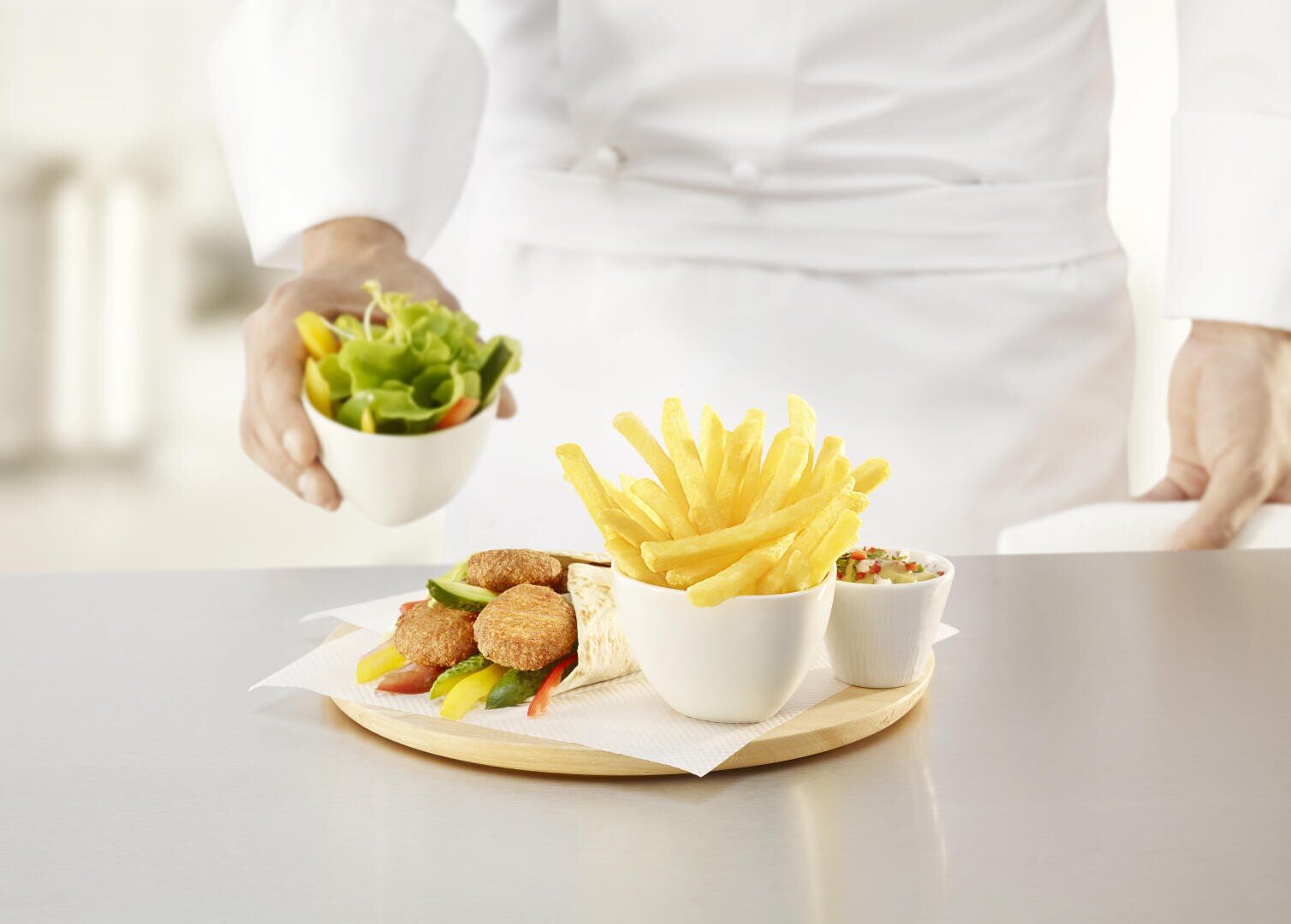 Un chef en uniforme blanc sert un plateau en bois contenant des frites, une salade, des boulettes de falafel, des bâtonnets de légumes et un petit bol de sauce sur un comptoir argenté.