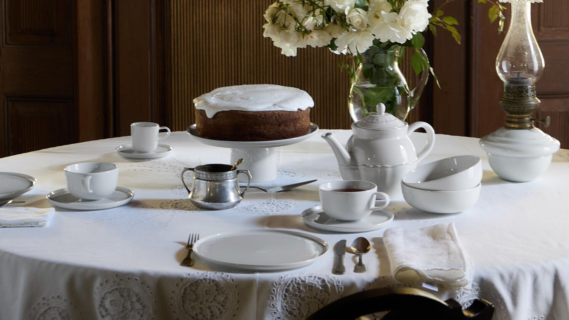 Une table ronde recouverte d'une nappe blanche est dressée pour le thé, avec un gâteau givré sur un support, une théière, des tasses et des soucoupes, des assiettes, des couverts, un sucrier et un vase de fleurs blanches.