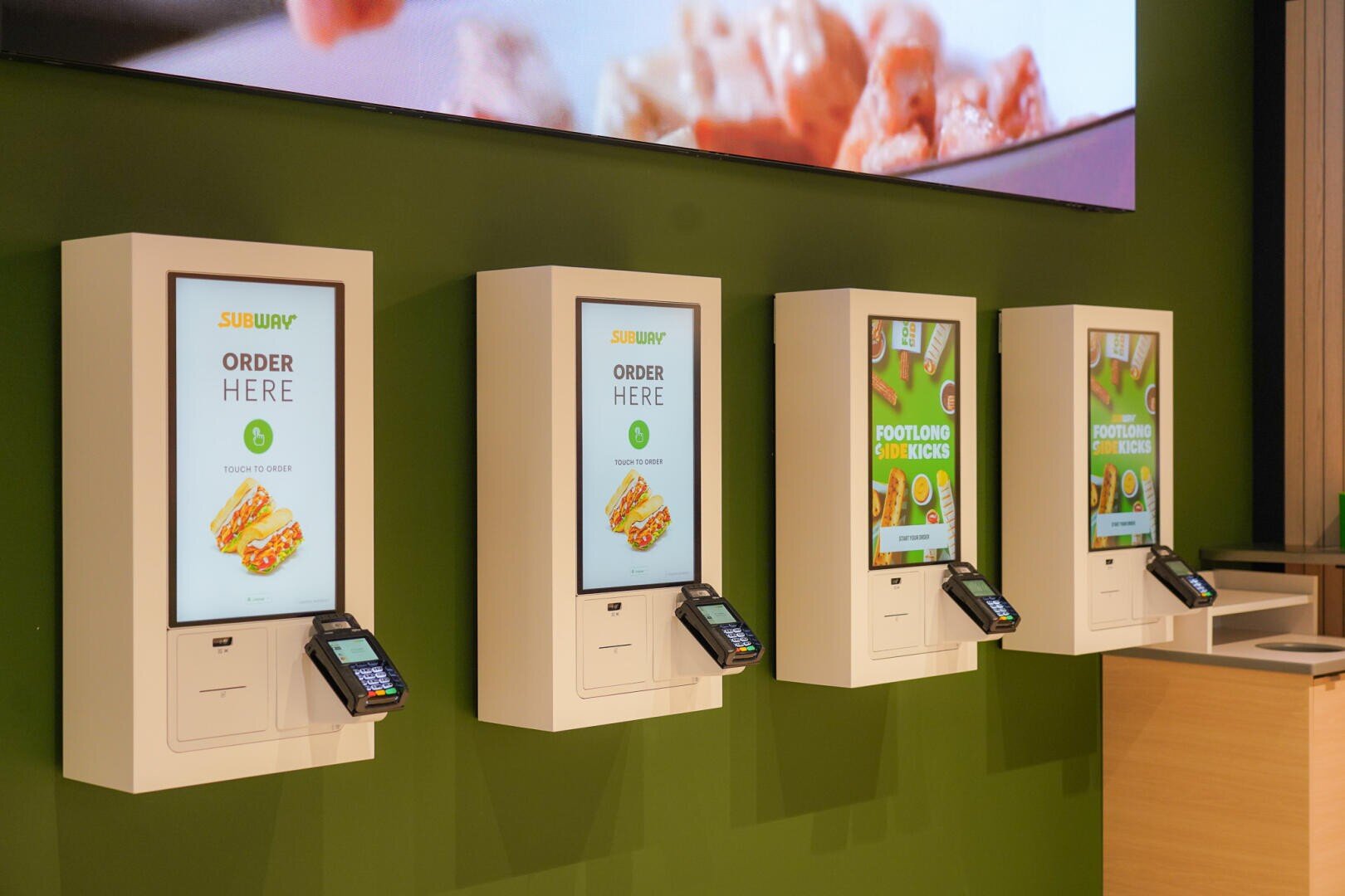 Vier Selbstbedienungs-Subway-Bestellkioske sind an einer grünen Wand angebracht, jeder mit einem Touchscreen, auf dem "Order Here" und Bilder von Sandwiches angezeigt werden. Unter jedem Bildschirm sind Kartenzahlungsterminals angebracht.