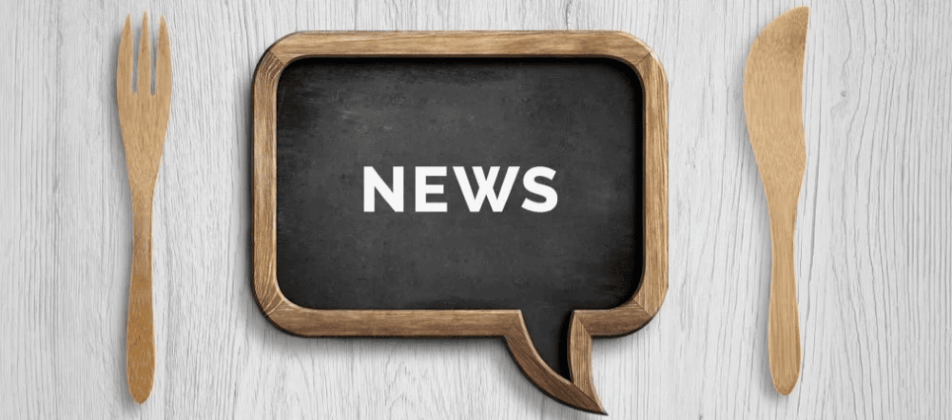 Auf einer Kreidetafel in Form einer Sprechblase steht in weißen Buchstaben das Wort NEWS, links eine Holzgabel und rechts ein Holzmesser, alles auf einer hellen Holzfläche.