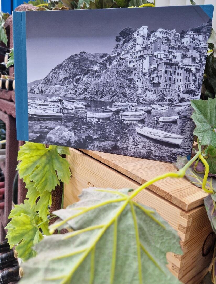 Ein Schwarz-Weiß-Fotobuch mit einem Bild eines Küstendorfes und Booten wird aufrecht auf einer Holzkiste präsentiert, umgeben von grünen Blättern und Pflanzen.