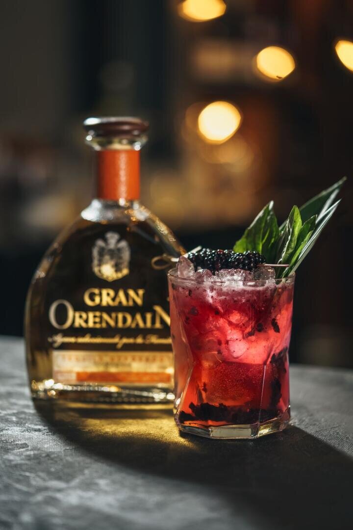 Ein Glas mit einem Beerencocktail mit Eis, frischen Brombeeren und grüner Garnitur steht vor einer Flasche Gran Orendain Tequila, mit warmen, verschwommenen Lichtern im Hintergrund.