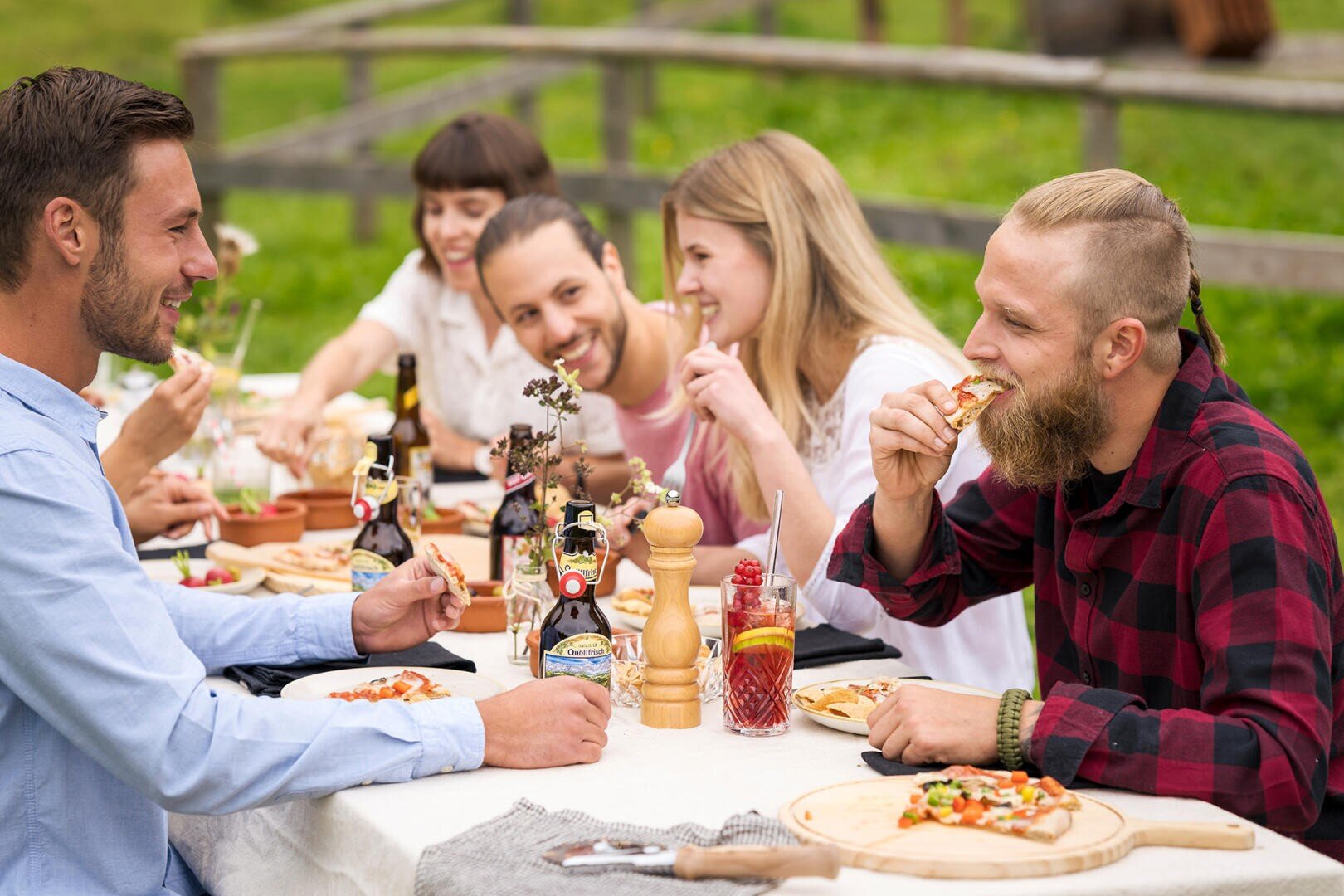 Eine Gruppe von Freunden sitzt draußen an einem Tisch, lächelt und genießt an einem sonnigen Tag gemeinsam eine Pizza. Getränke und Gewürze stehen auf dem Tisch, und im Hintergrund ist Grün zu sehen.