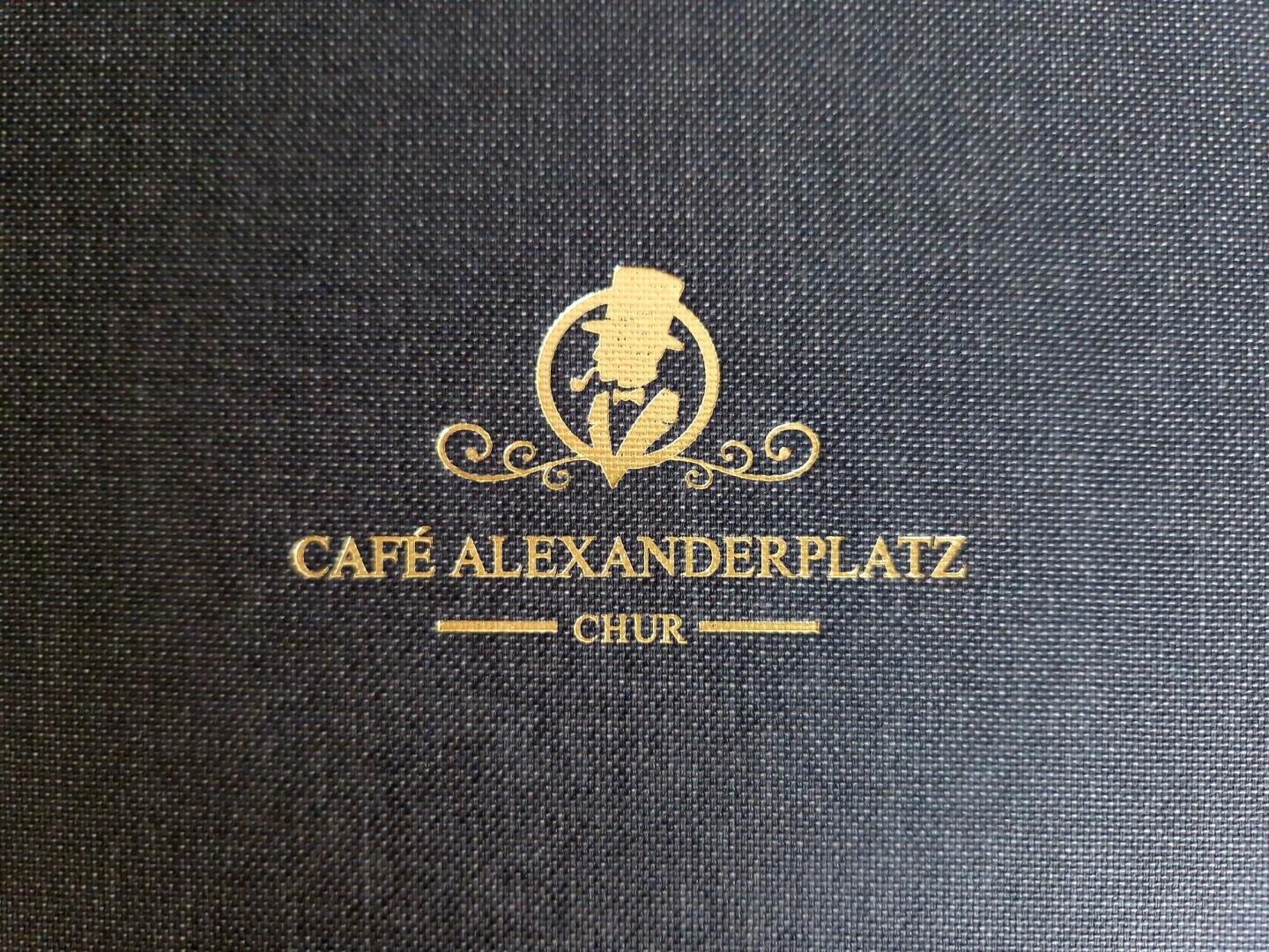 Schwarze, strukturierte Oberfläche mit einem goldenen Logo einer Person mit Zylinder und Stock, über dem Text CAFÉ ALEXANDERPLATZ CHUR in eleganter Schrift, mit dekorativen Wirbeln um das Logo.