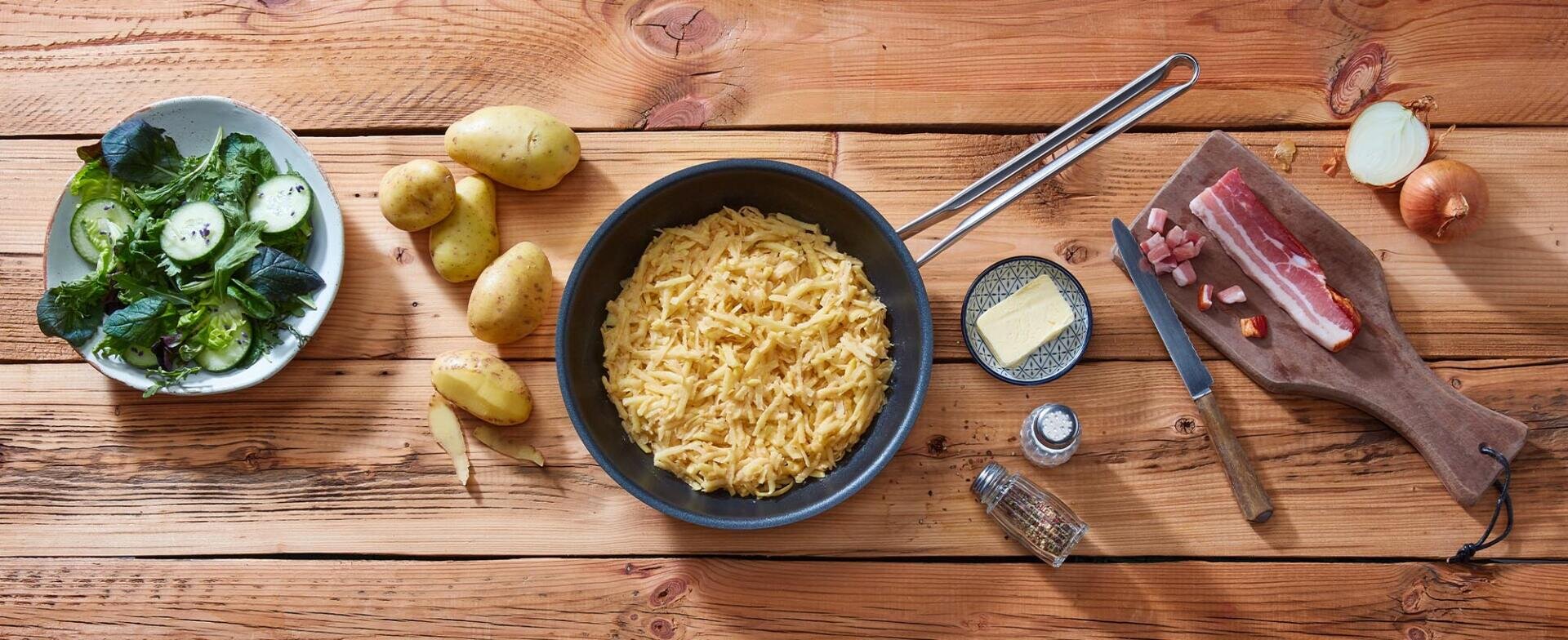 Eine Pfanne mit gekochten geriebenen Kartoffeln steht auf einem Holztisch, umgeben von rohen Kartoffeln, Butter, Salat, Speck, gehackten Zwiebeln, einem Messer und Gewürzen auf einem Schneidebrett.