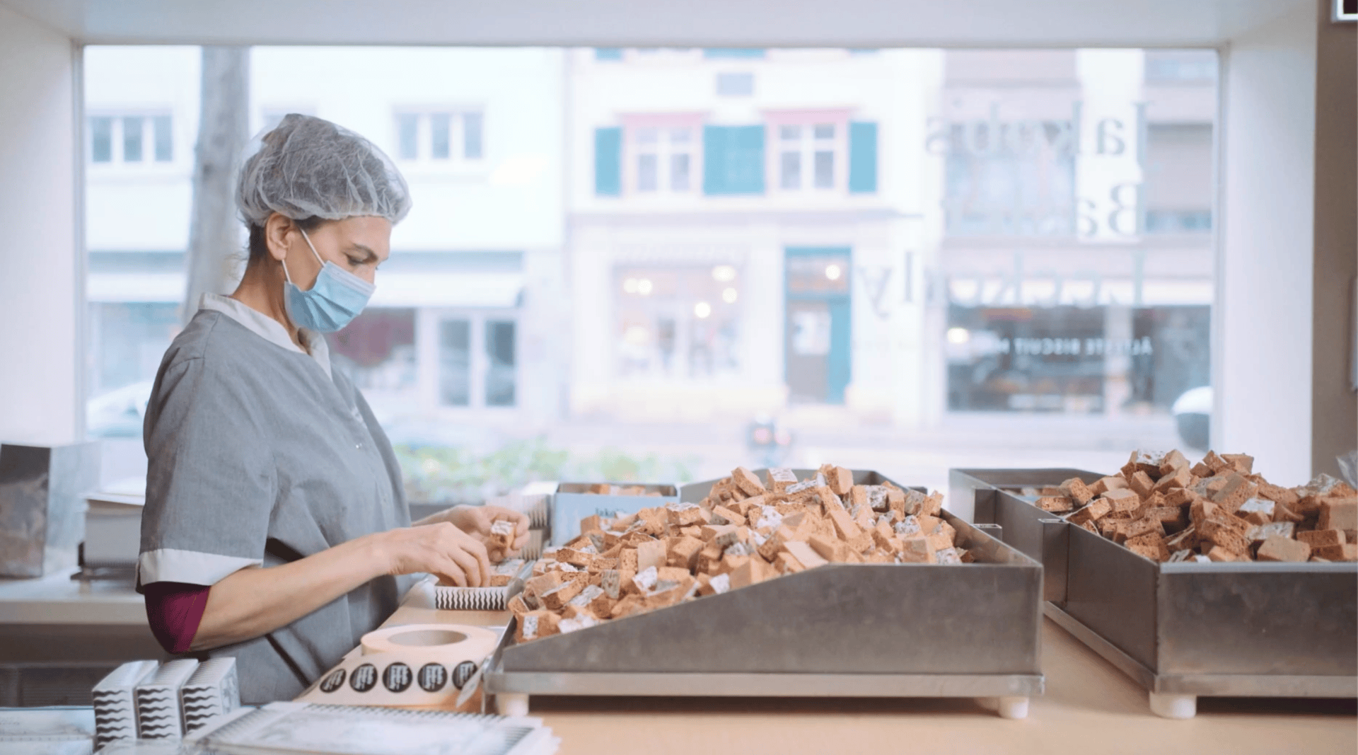 Une femme portant une charlotte, un masque et un uniforme gris trie des bouchons dans des plateaux à un poste de travail dans une pièce lumineuse avec de grandes fenêtres montrant des bâtiments à l'extérieur.