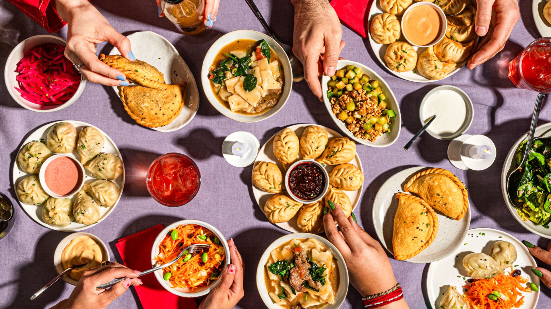 Une table vivante avec différentes boulettes, pâtisseries, plats de légumes, sauces et boissons. Plusieurs mains se donnent la main pour partager et servir la nourriture, créant ainsi une scène de repas animée et communautaire.